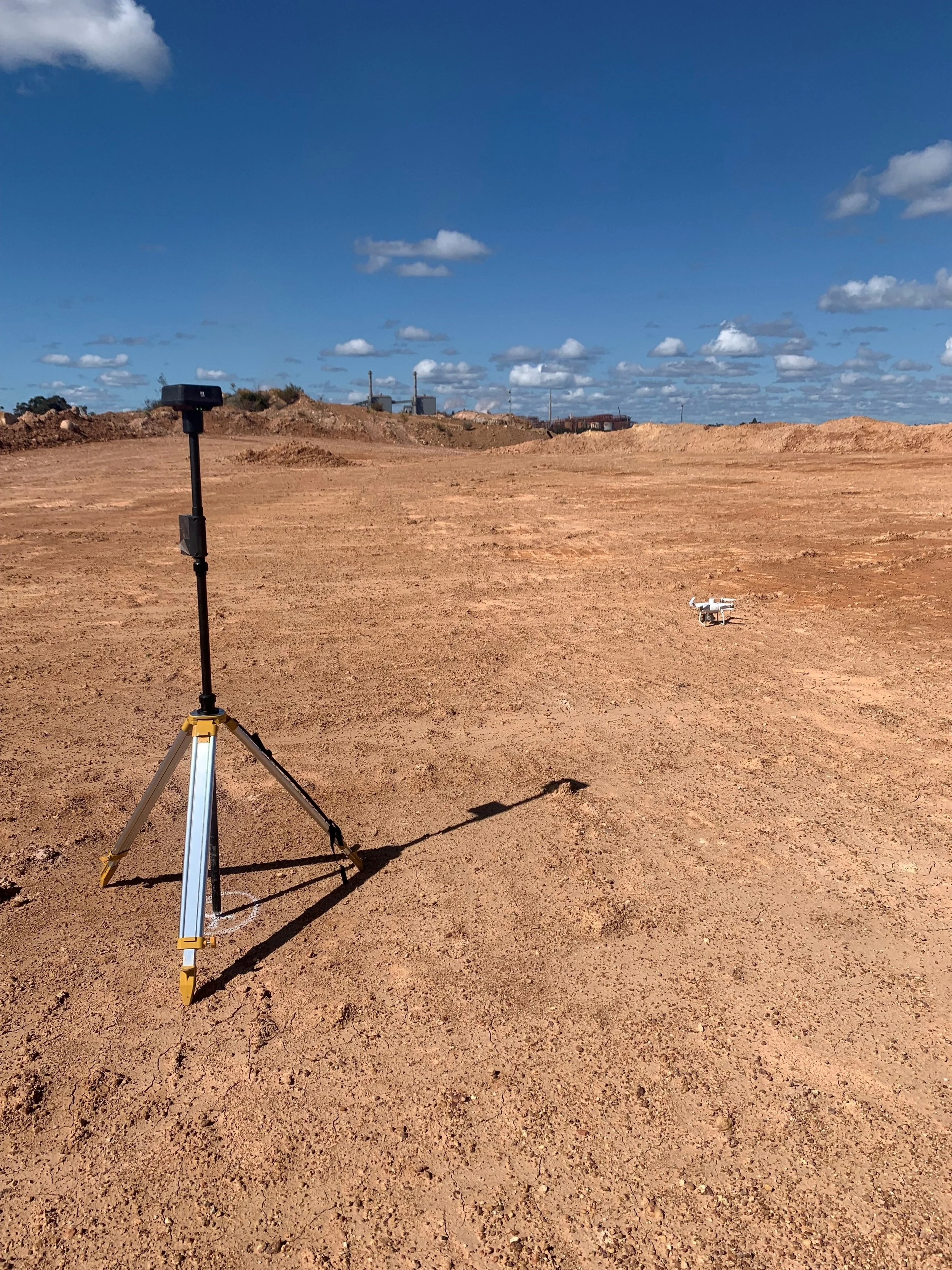 A drone flying over a flat, sandy open area with a camera mounted on a tripod in the foreground. There are some buildings and structures in the background under a partly cloudy sky.