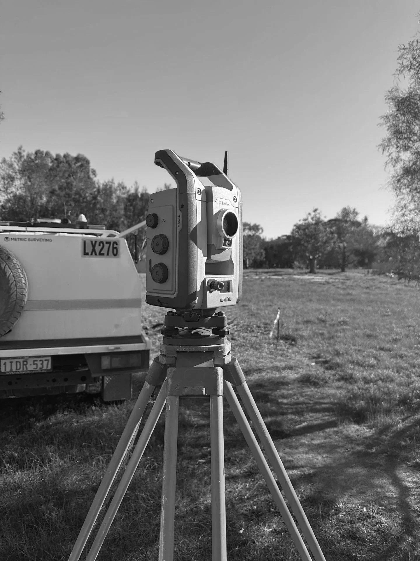 A surveying instrument mounted on a tripod outdoors with a survey truck and trees in the background.