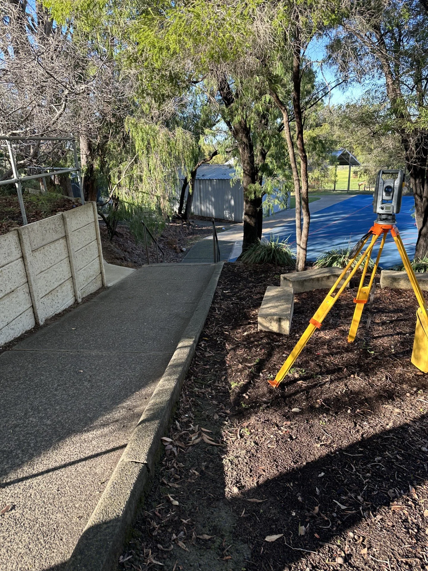 A construction site on a small outdoor slope with a newly paved sidewalk, a tripod-mounted surveying instrument, trees, a grassy area, and a distant shed, under a clear blue sky.