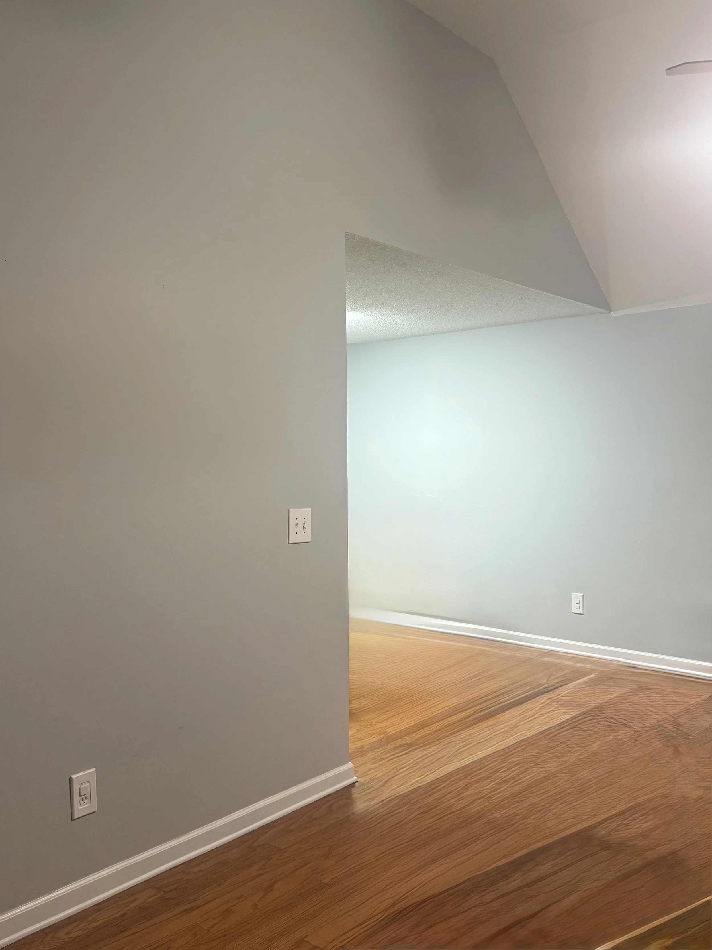 Empty room with light gray walls, hardwood flooring, and a sloped ceiling. There are electrical outlets on the walls.
