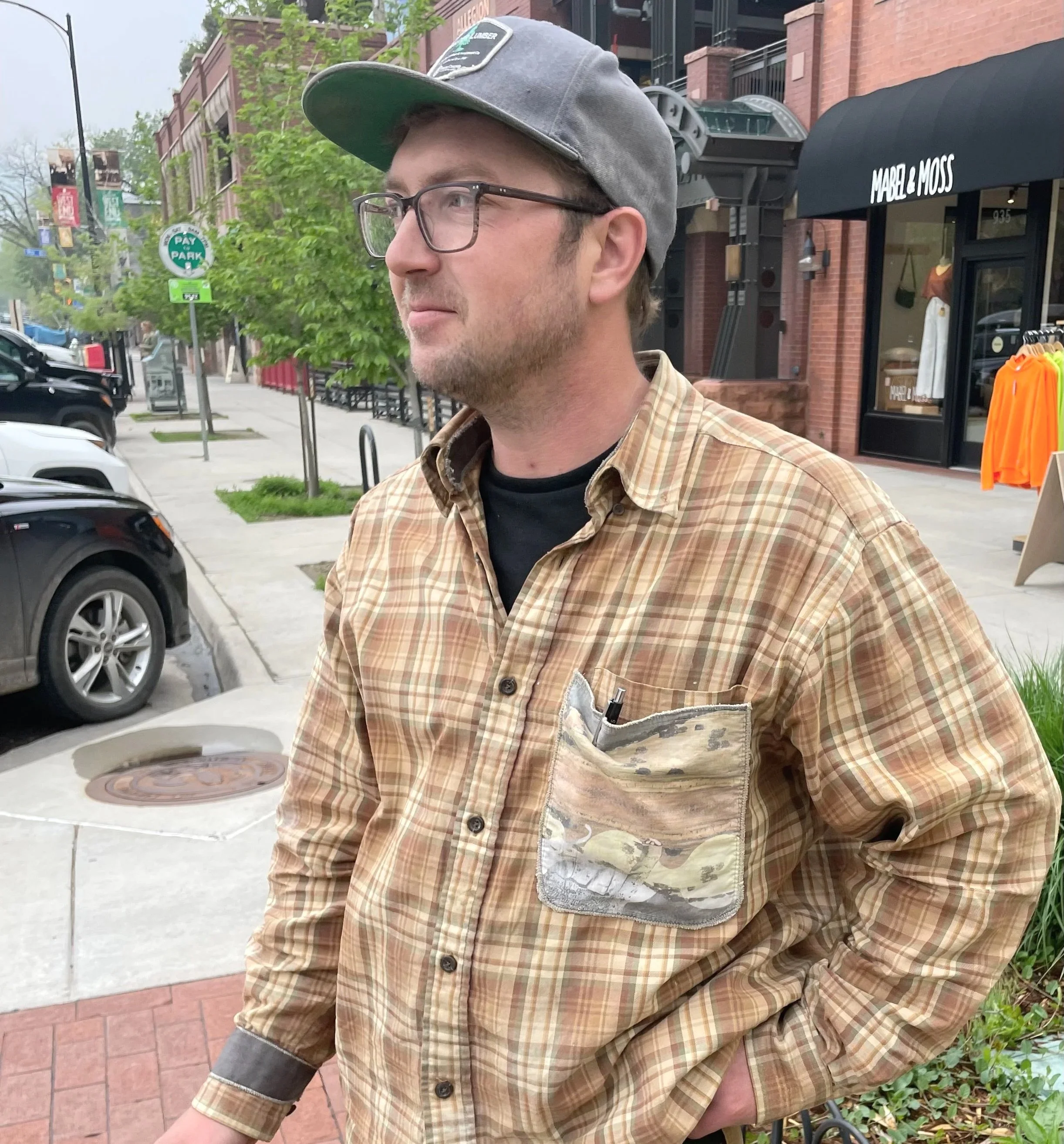 A man standing on a sidewalk in front of shops, wearing glasses, a gray cap, a plaid shirt over a black t-shirt, with pens in his shirt pocket, and looking to the side.