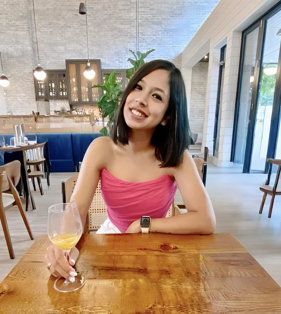 A smiling young woman with black shoulder-length hair, wearing a pink strapless top, sits at a wooden table in a modern restaurant. She has a smartwatch on her left wrist and is holding a glass of white wine with lemon slices. The restaurant has a light-colored brick wall, hanging pendant lights, large windows, and some green plants.