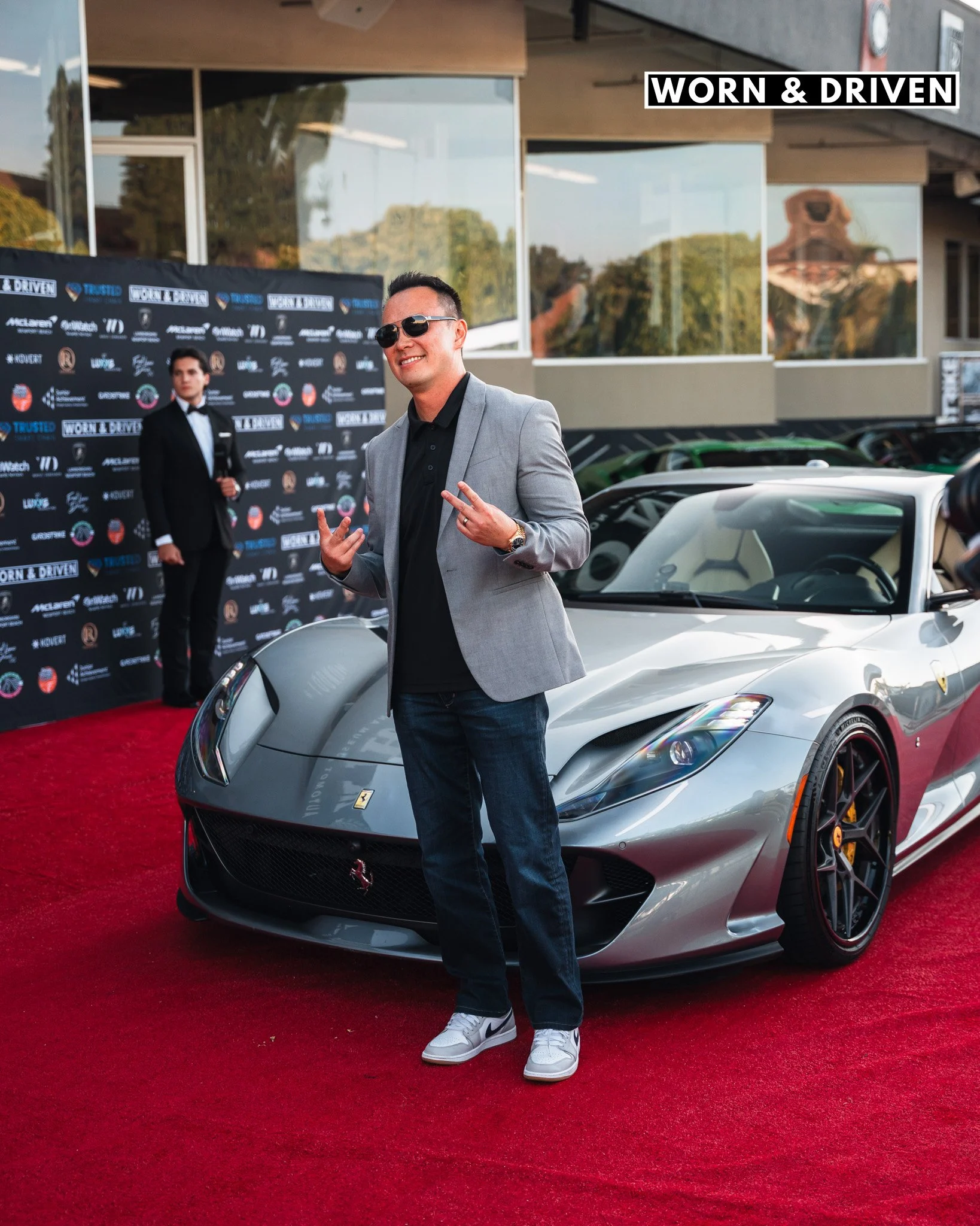A man wearing sunglasses, a gray blazer, black shirt, and jeans standing on a red carpet in front of a silver sports car at an outdoor event, with people and a backdrop with various logos behind him.