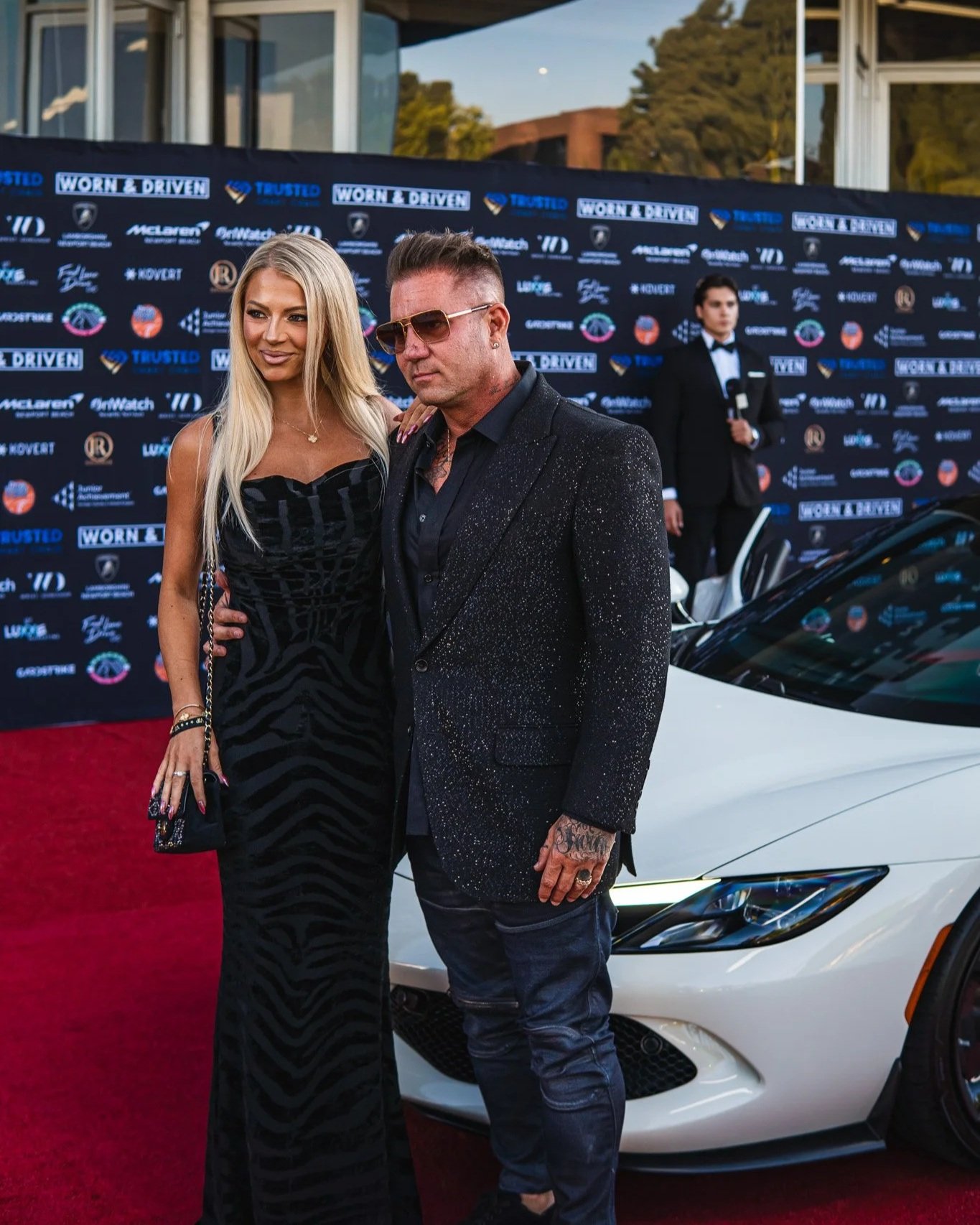A man and woman pose together on a red carpet at a formal event, with a backdrop featuring various logos. The woman is wearing a black dress with a pattern, and the man is dressed in a black speckled blazer and sunglasses. They are standing in front of a white car, and another person in a tuxedo is visible in the background.