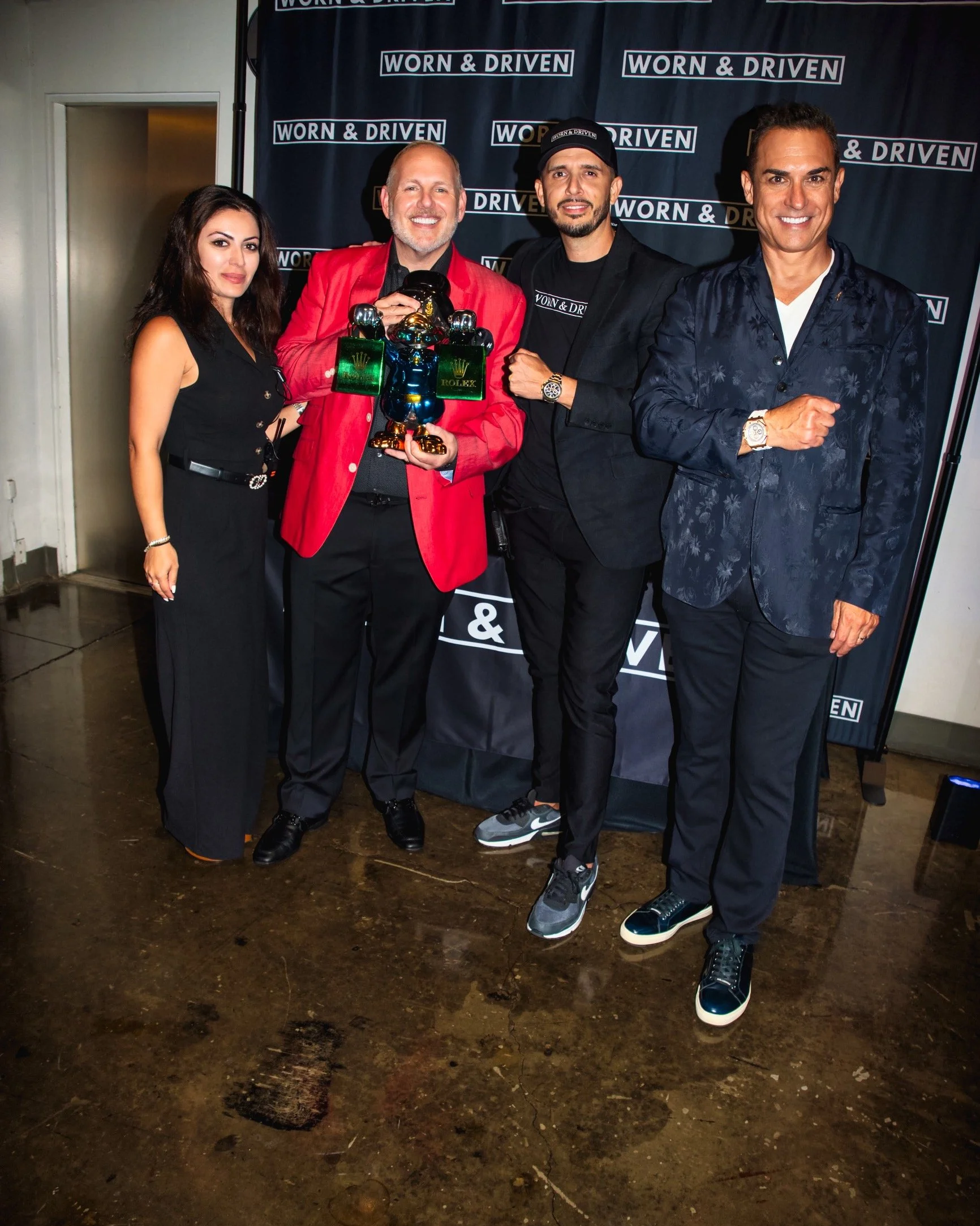 Four people standing in front of a black backdrop with the words 'Worn & Driven' repeated. The person second from the left is holding a trophy with a gold crown and assorted metallic objects, wearing a red jacket. The others are dressed in stylish attire, all smiling at the camera.