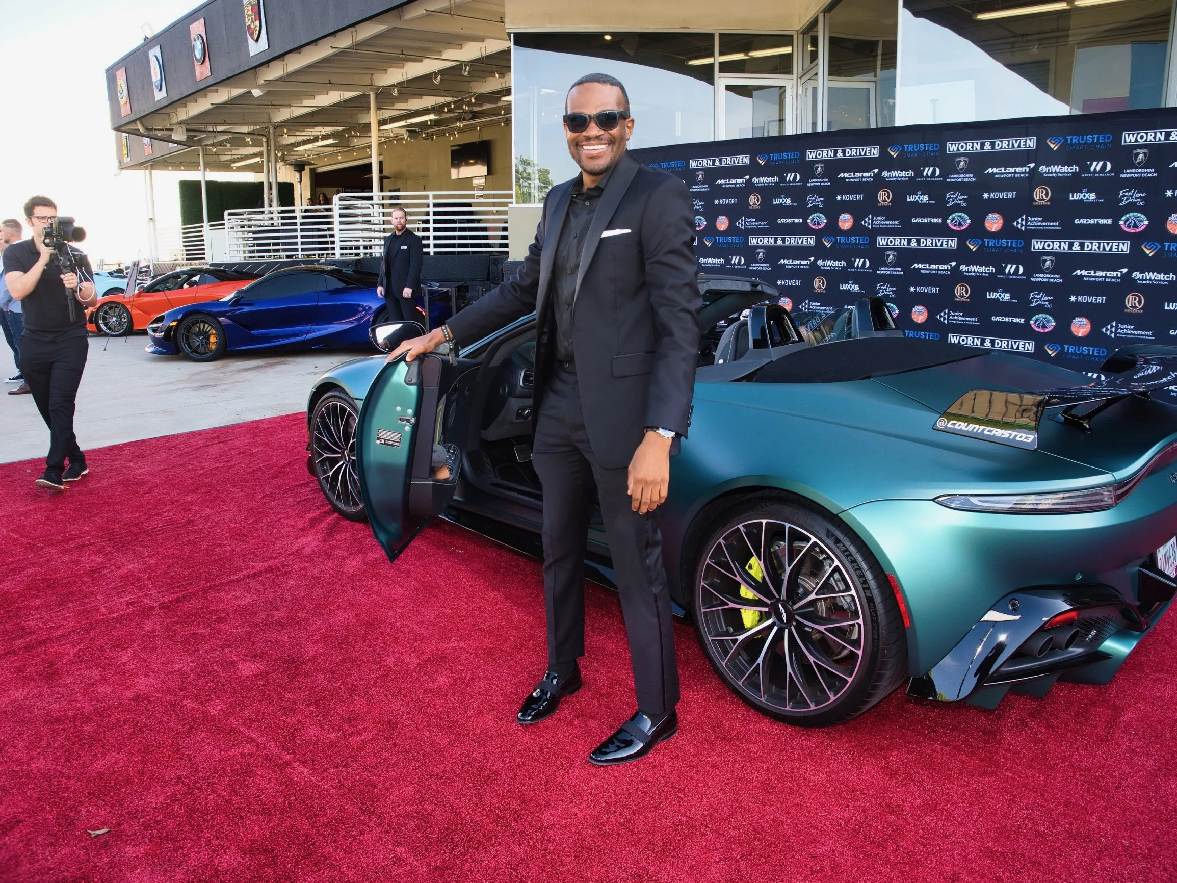 Man in black suit with sunglasses smiling standing next to a green sports car with open door, at an event with luxury cars and a branded backdrop.