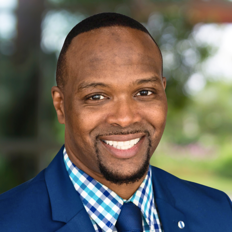 Portrait of a smiling man wearing a blue suit, checkered shirt, and blue tie outdoors.