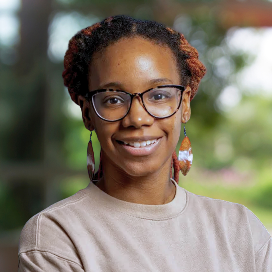 A smiling woman with glasses, earrings, and a beige shirt smiling outdoors with a blurred natural background.