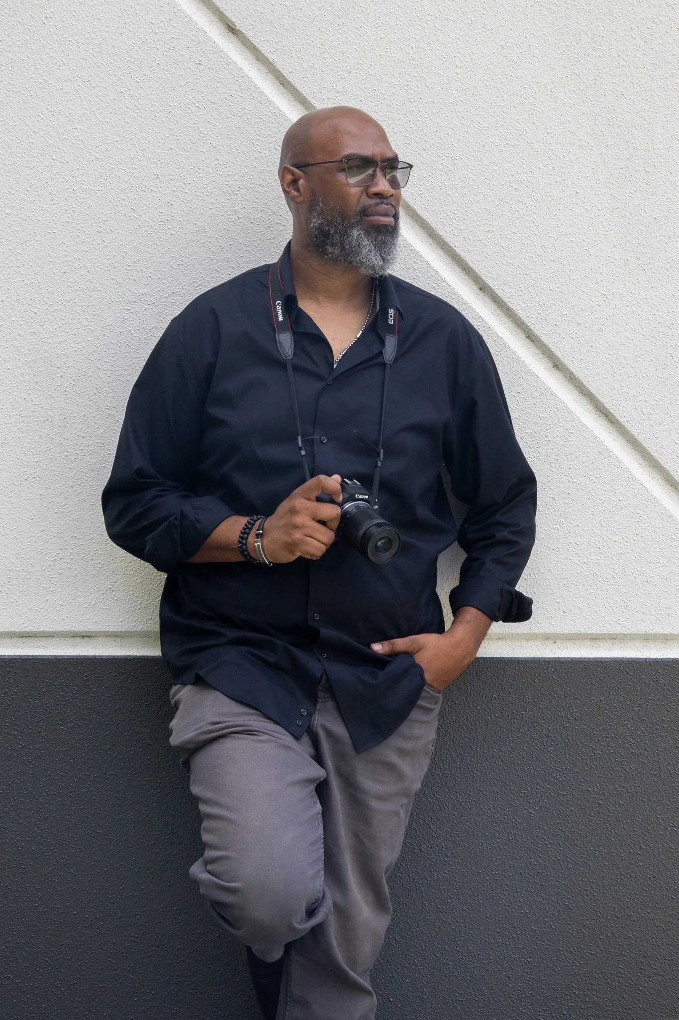Warren Miles of Warm Lens Visions wearing a black shirt stands against a white wall with dark gray lower panel, holding a camera around his neck and looking to the side.