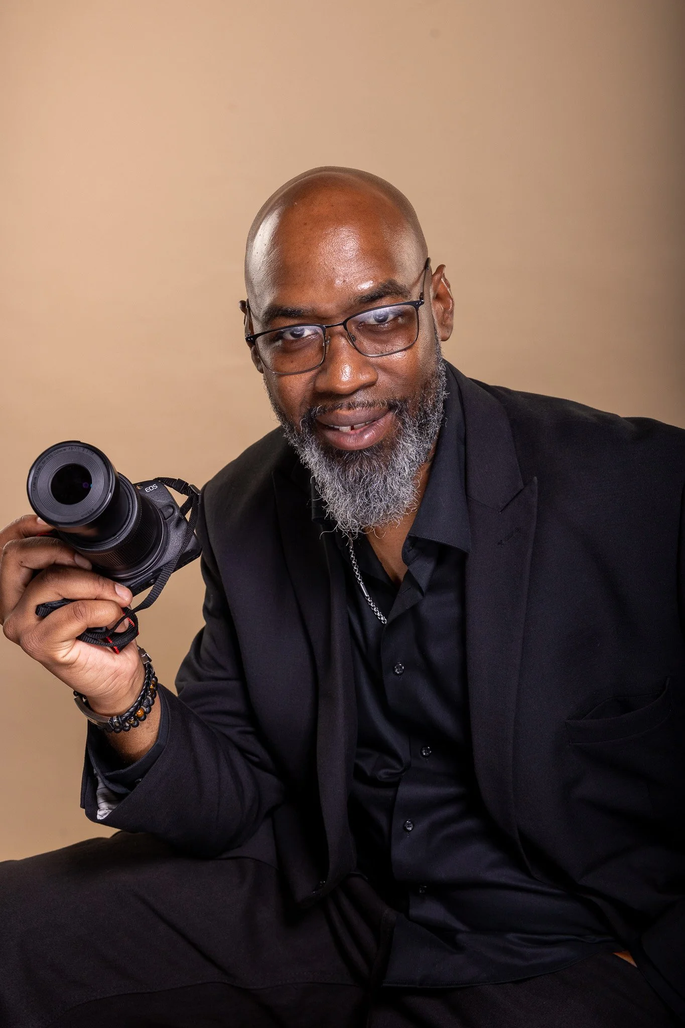 Warren Miles of Warm Lens Visions wearing a black suit and shirt, holding a camera in his right hand, against a plain beige background.