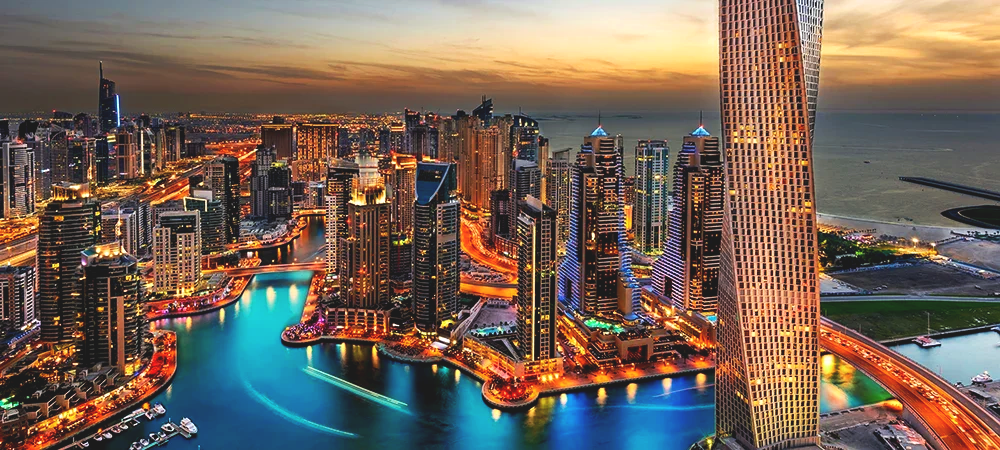 Aerial view of a brightly lit modern city skyline at sunset, with tall skyscrapers, a winding river, and a large twisting skyscraper in the foreground in Dubai, United Arab Emirates.