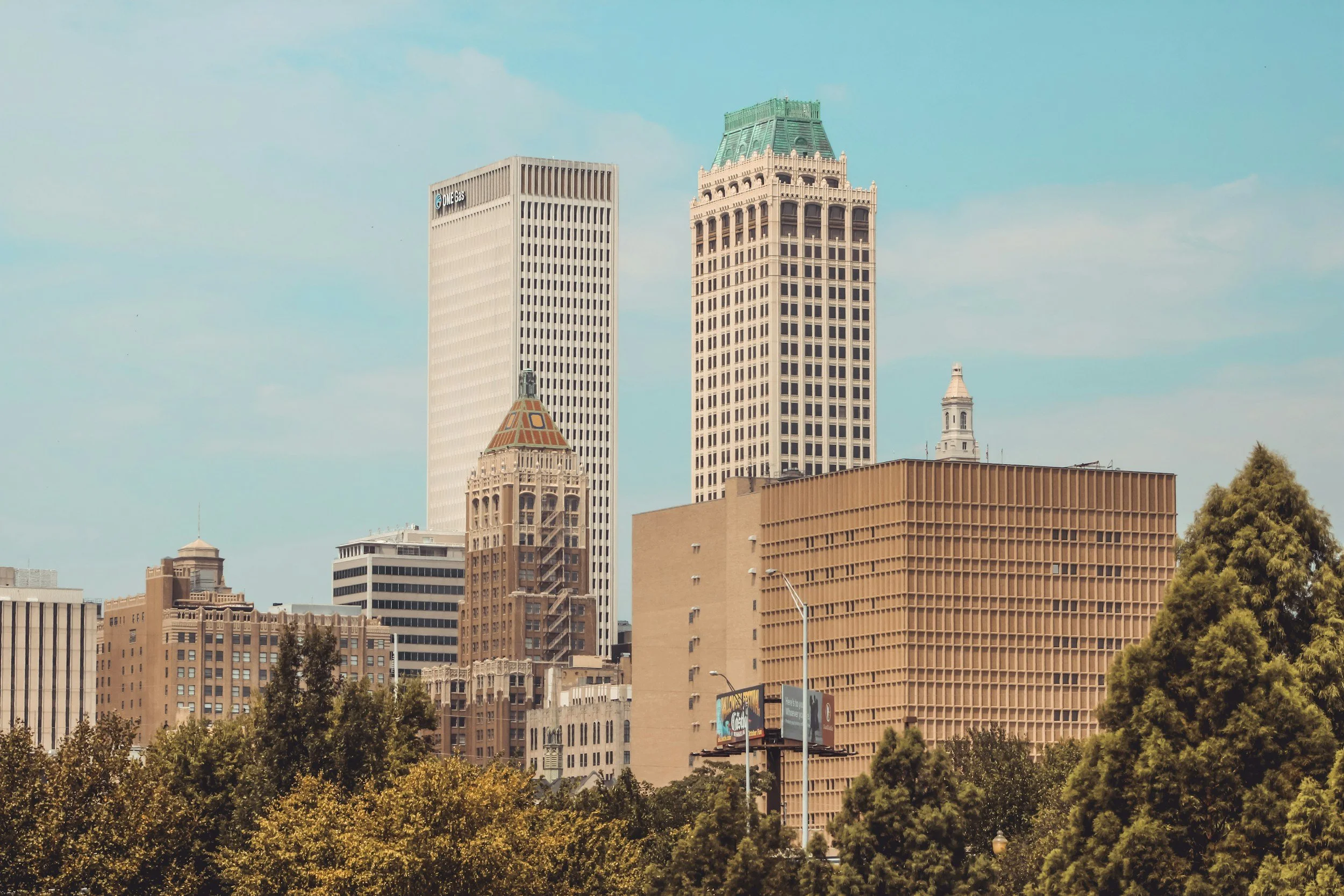 Cityscape with tall buildings and trees in the foreground, under a partly cloudy sky.