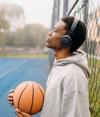 Young man wearing headphones holding a basketball on a sports court near a chain-link fence.
