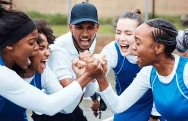 A group of women and a coach engaging in a friendly arm-wrestling match, smiling and celebrating on a sports field.