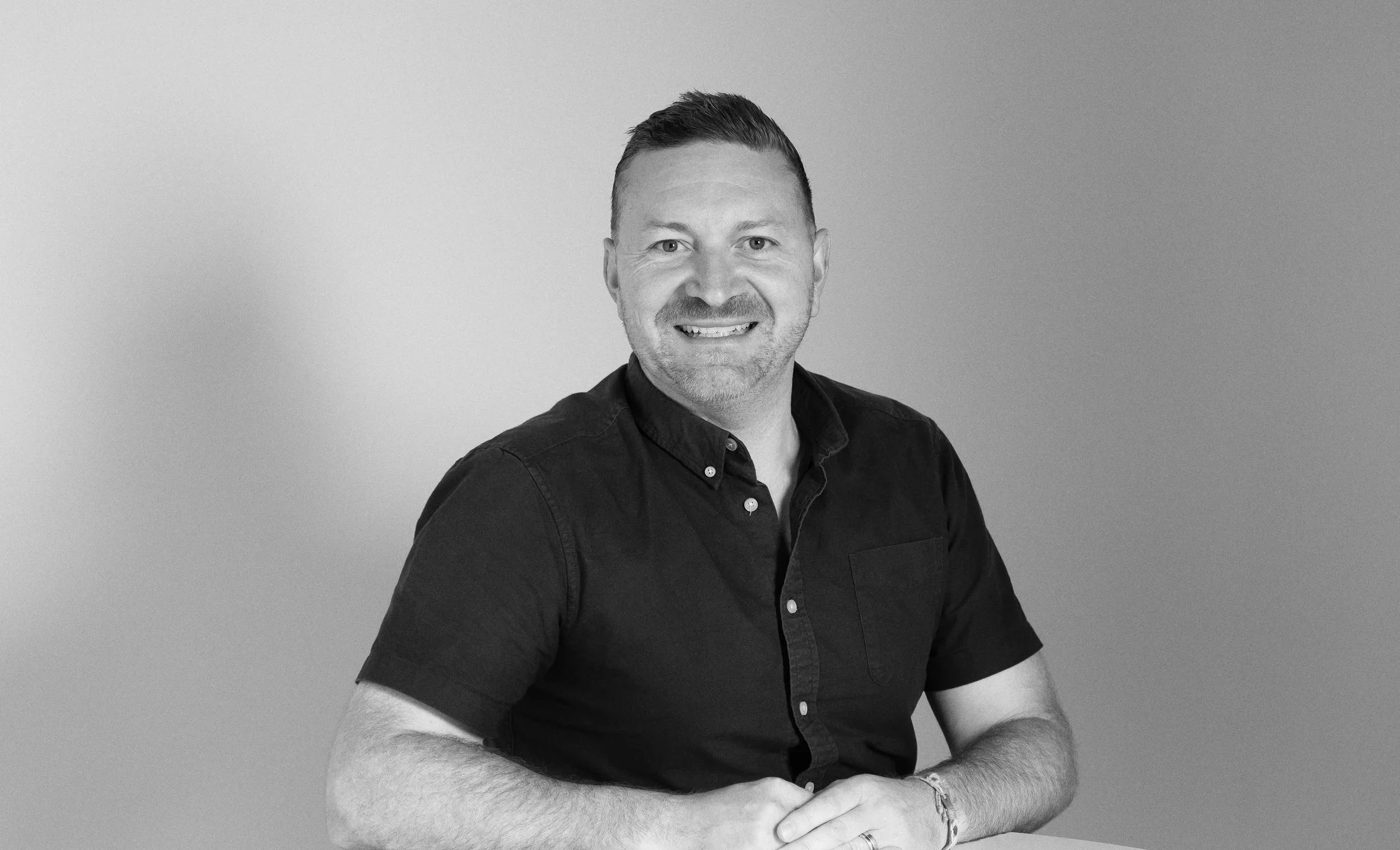 Black and white portrait of a man in a checkered blazer and black shirt, smiling with hands folded on a table against a plain background.