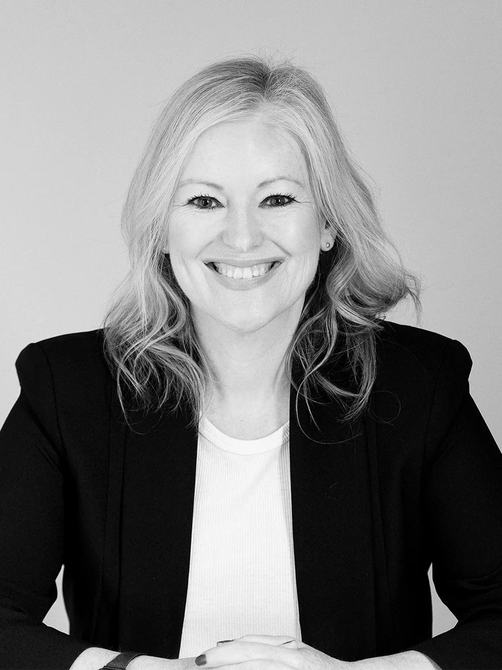 Black and white portrait of a smiling woman with shoulder-length wavy hair, wearing a dark blazer over a light top, standing against a plain light background.