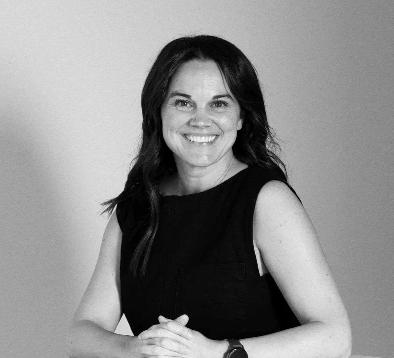 Black and white photo of a woman with shoulder-length dark hair, smiling, wearing a white blazer over a dark buttoned top, standing against a plain light background.