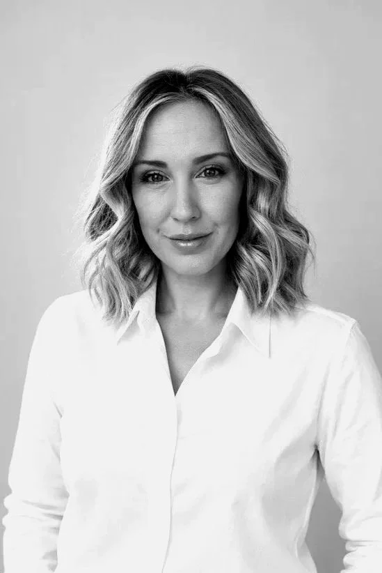 Black and white photo of a woman with wavy, shoulder-length hair wearing a white button-up shirt, smiling at the camera against a plain background.