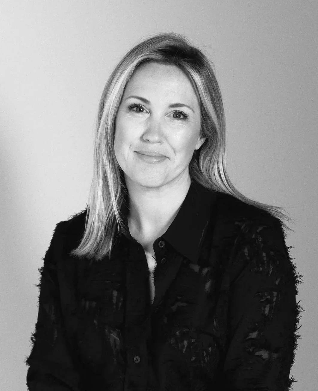 Black and white photo of a woman with wavy, shoulder-length hair wearing a white button-up shirt, smiling at the camera against a plain background.