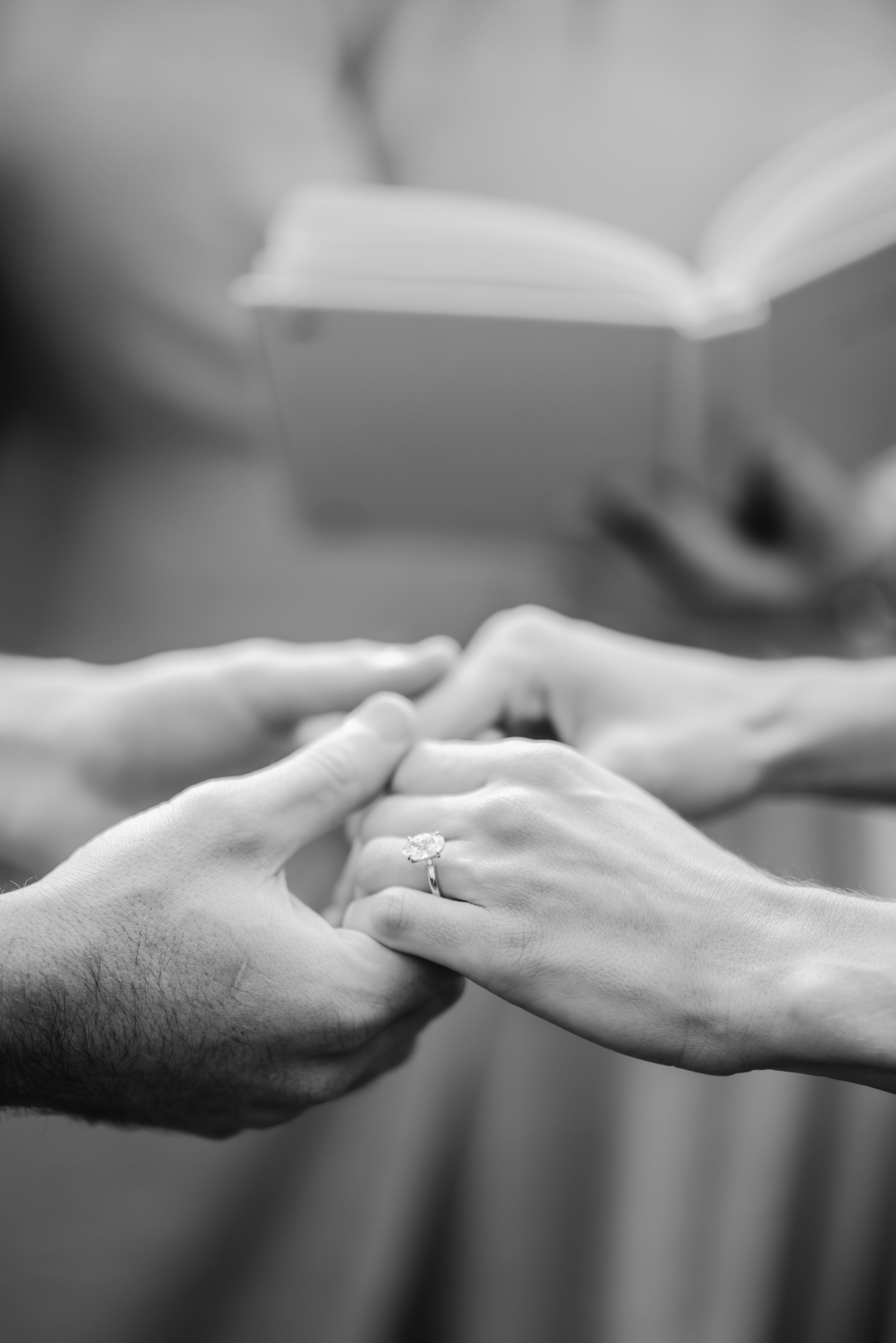 Close-up of a couple holding hands during a wedding ceremony, with an engagement ring visible on the woman's finger. Background includes an open book and blurred objects.