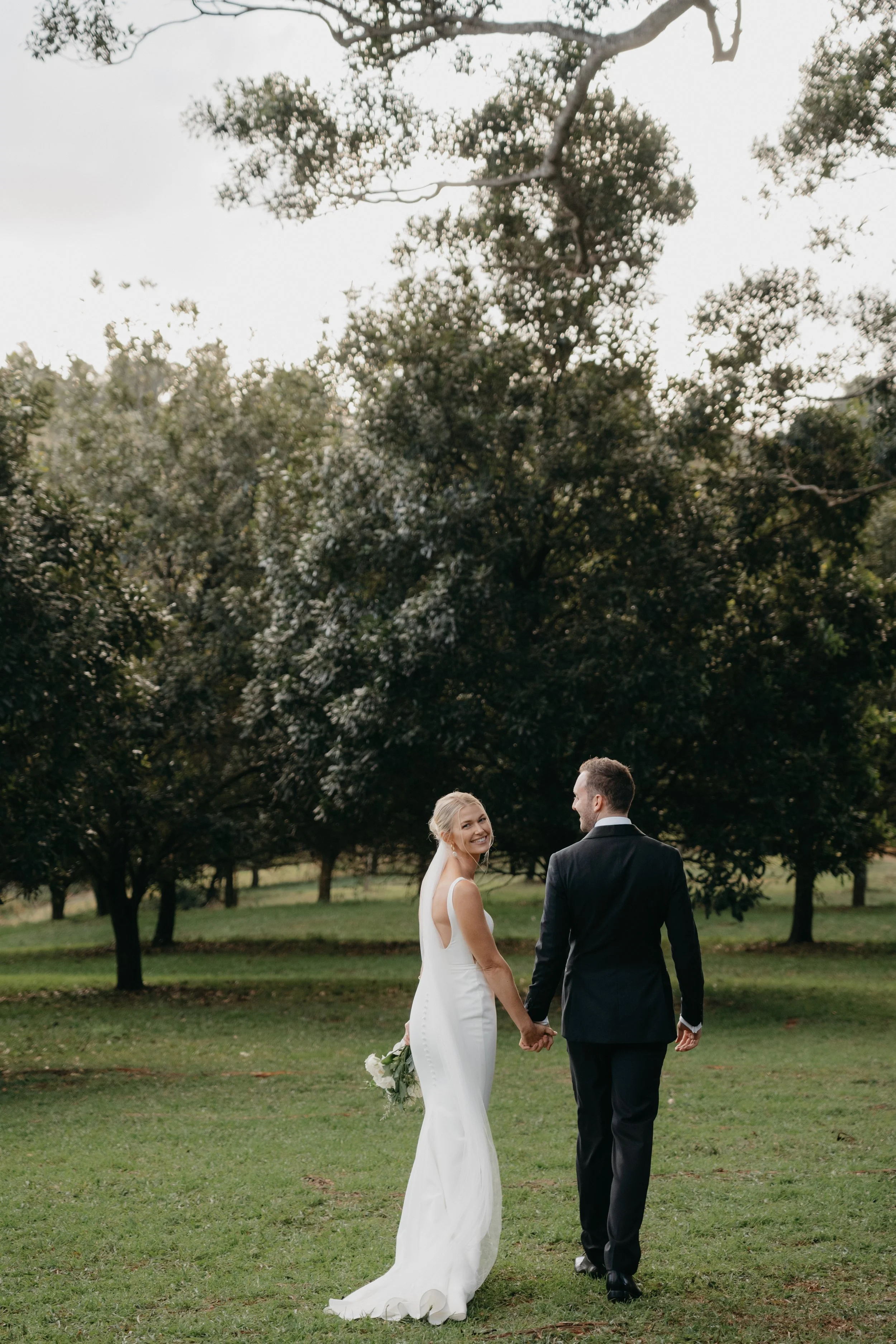 A bride and groom holding hands and walking after their wedding ceremony with their marriage celebrant. The bride is smiling and wearing a white wedding dress, the groom is dressed in a black suit and tie.