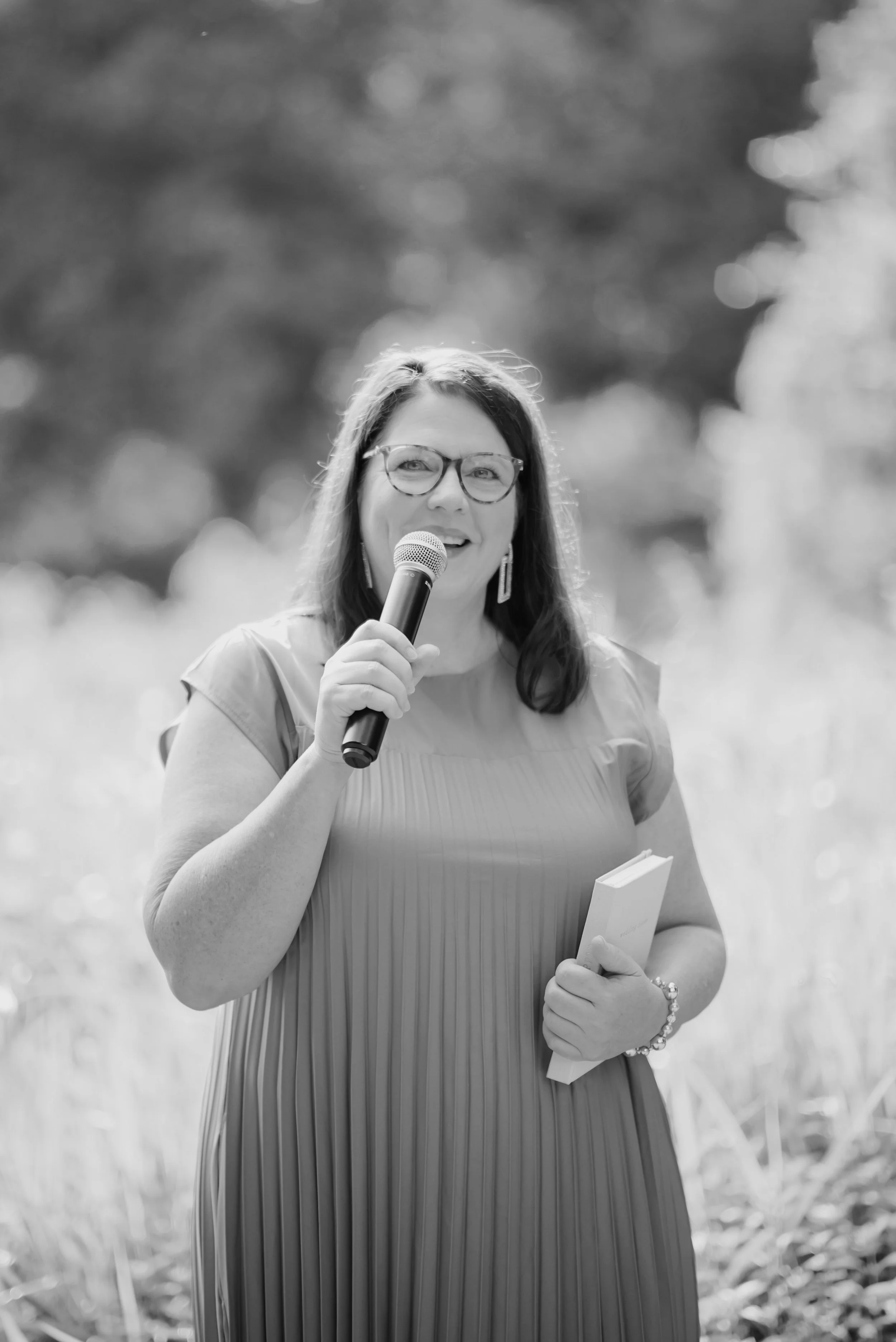 A marriage celebrant delivering a wedding ceremony. Holding a microphone, smiling, and looking at the bride and groom and wedding guests.