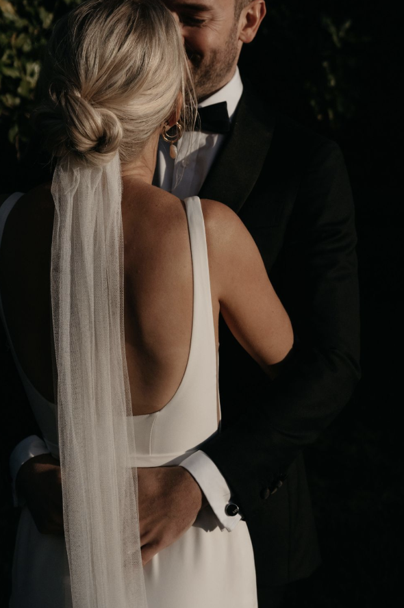 A bride and groom share an intimate moment at their wedding ceremony with their marriage celebrant. The bride's back is to the photographer  and her veil is flowing down her back.