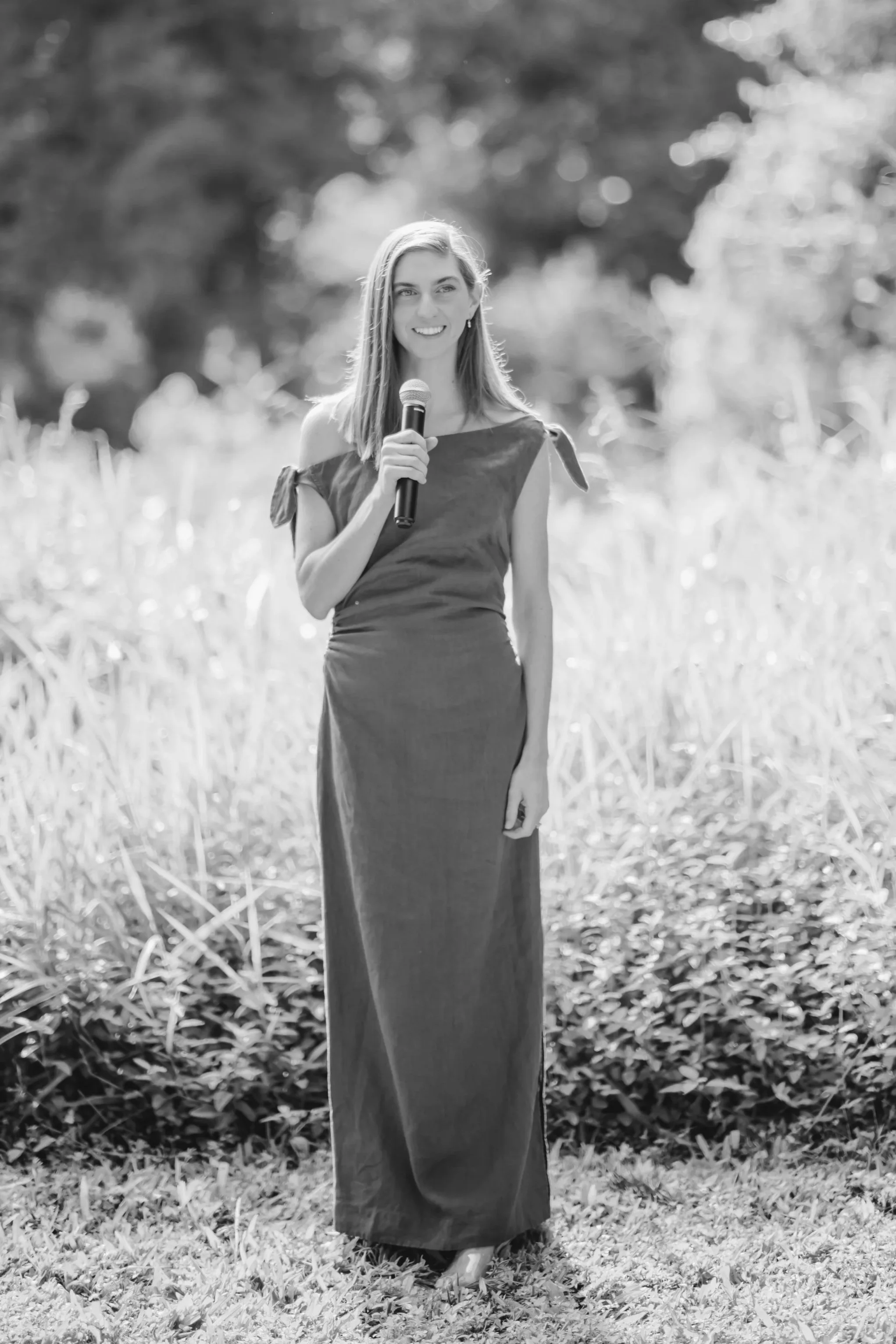 A marriage celebrant in a long dress delivering a wedding ceremony. Holding a microphone, smiling, and looking at the bride and groom and wedding guests.