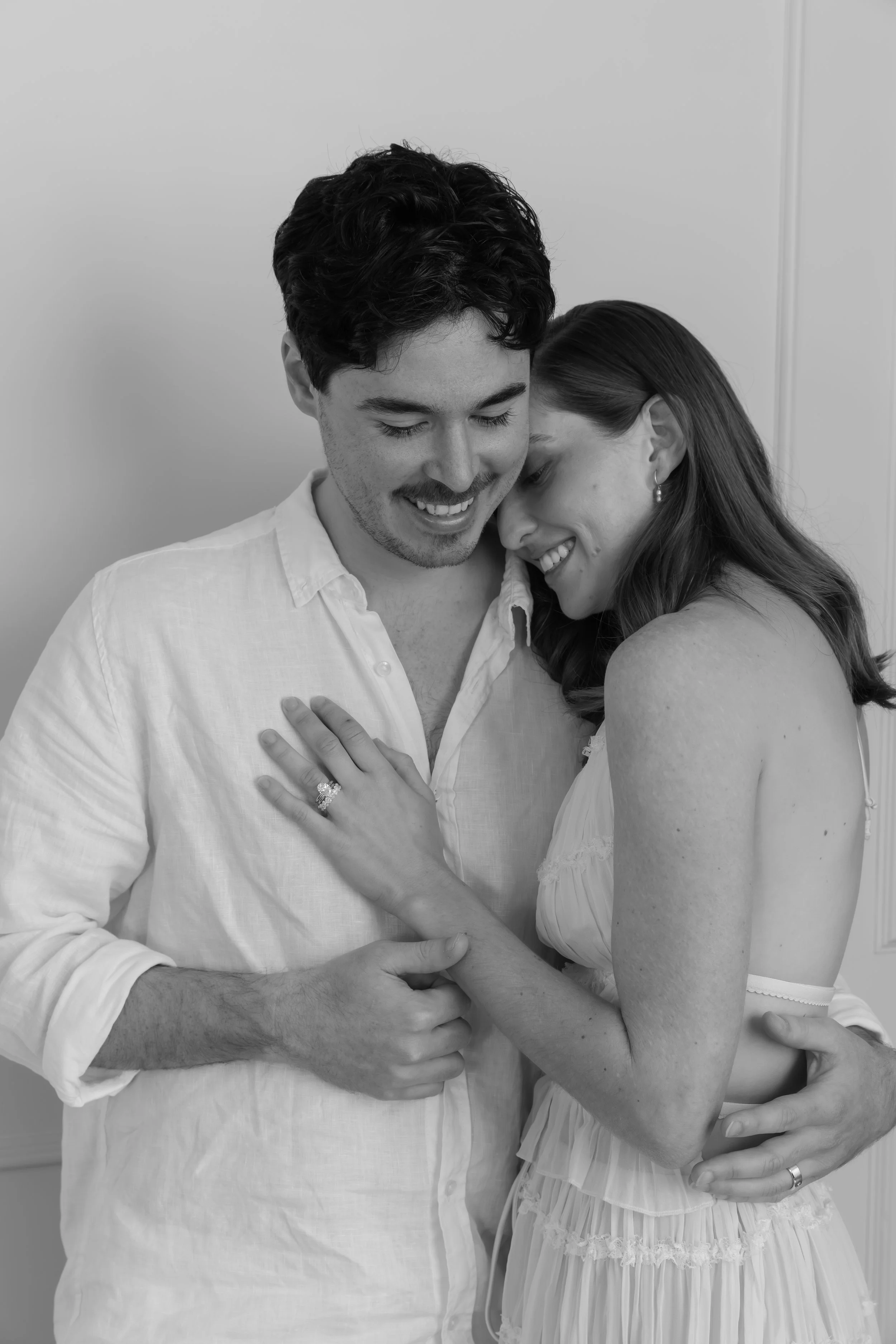 Black and white photo of a smiling couple in an embrace. The woman is touching the man's chest with her left hand, displaying an engagement ring, and her right arm wrapping around his waist. The man has his right hand on the woman's waist, and they are both looking down with happy expressions.