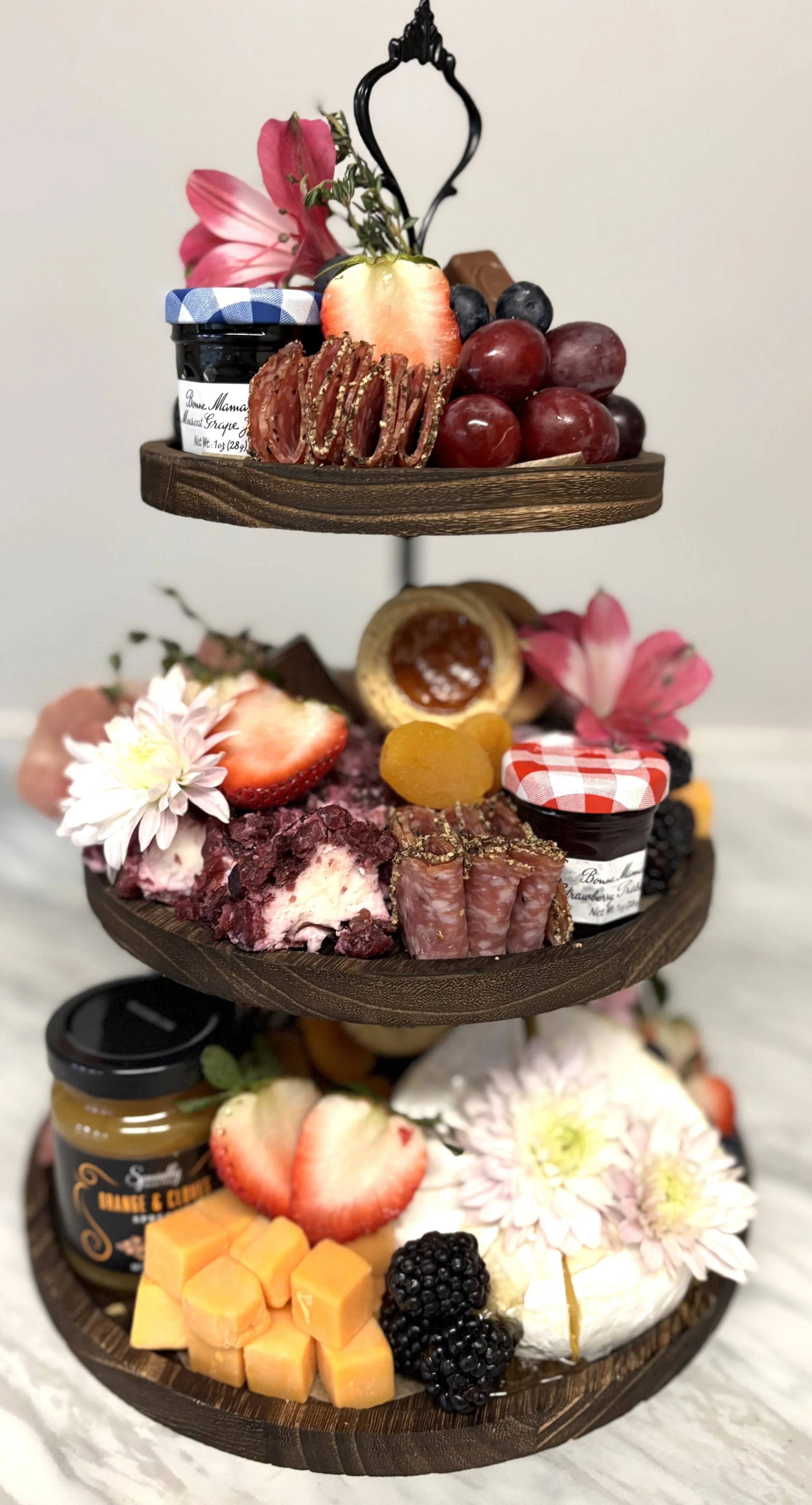 Three-tiered wooden serving tray filled with various desserts, fruits, and flowers. The top tier has grapes, blueberries, a strawberry, pink flowers, and a small jar of jam. The middle tier contains more strawberries, dried apricots, a tart, and a jar of berry jam. The bottom tier features cubed cheese, sliced strawberries, blackberries, flowers, and a jar of caramel.