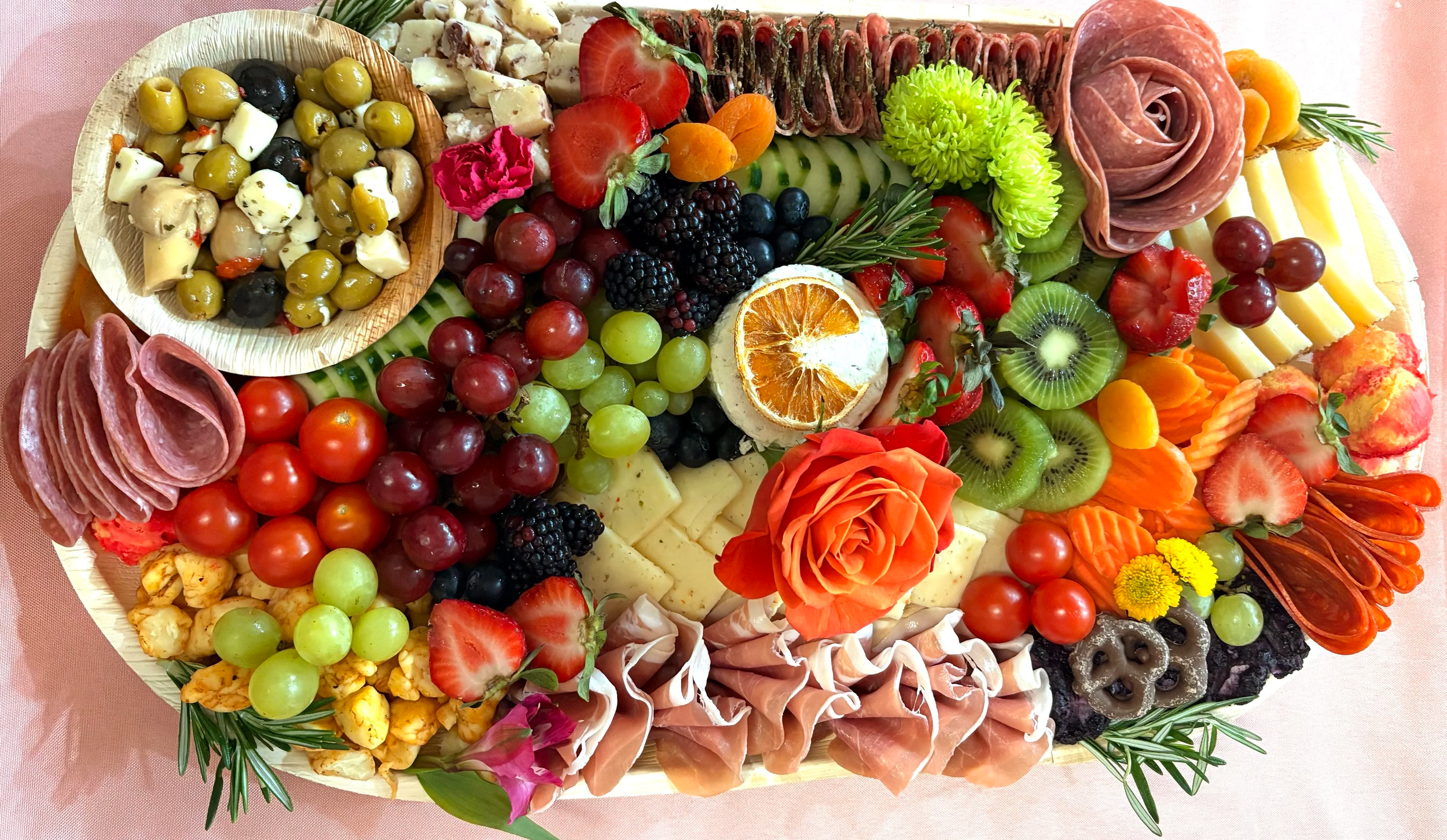 A platter of assorted cheeses, fresh fruits including strawberries, grapes, kiwi, and blackberries, cured meats, olives, crackers, and garnishes such as flowers and herbs.