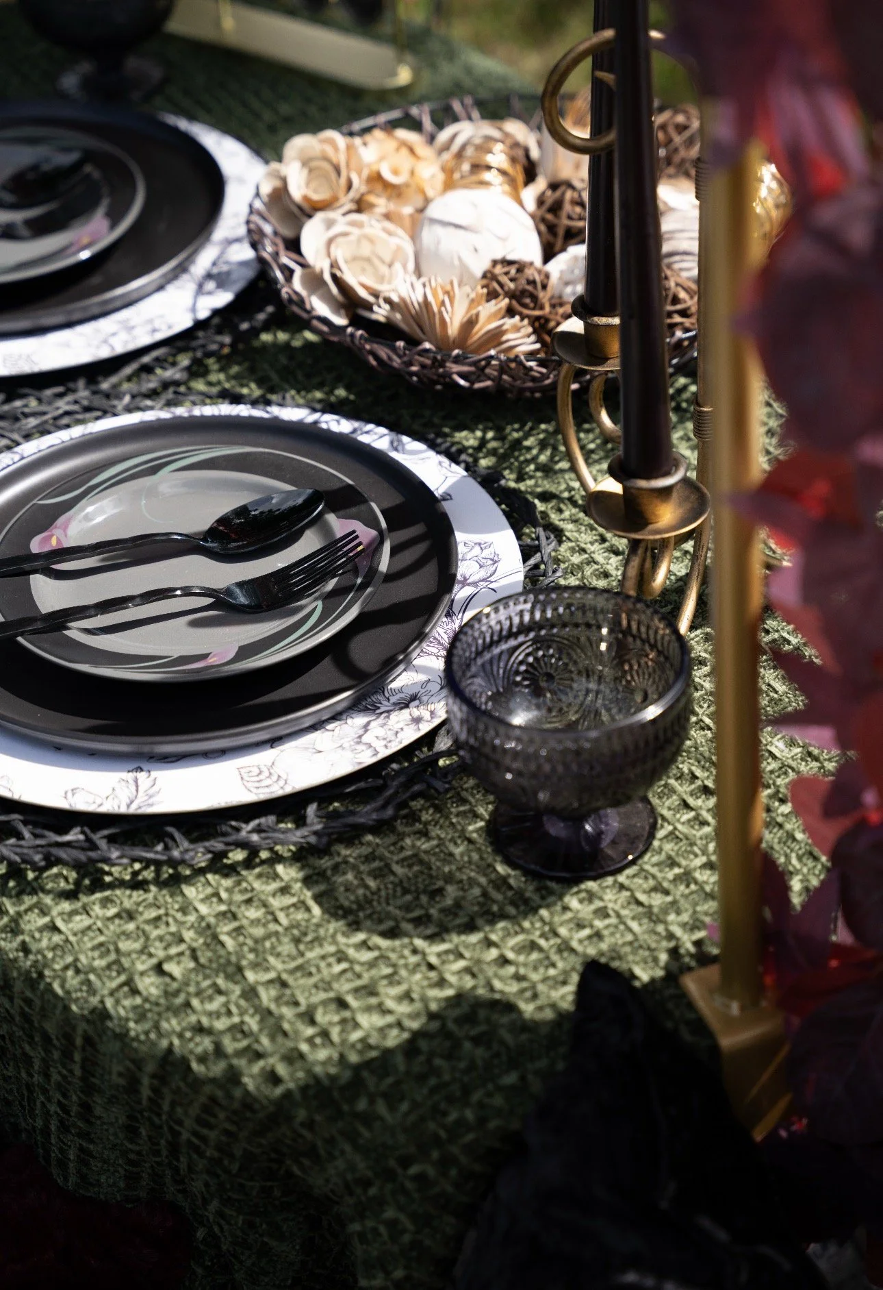 Outdoor table setting with black and white floral dinnerware, a black spoon and fork, a glass goblet, and decorative items including a basket of flowers and candles.