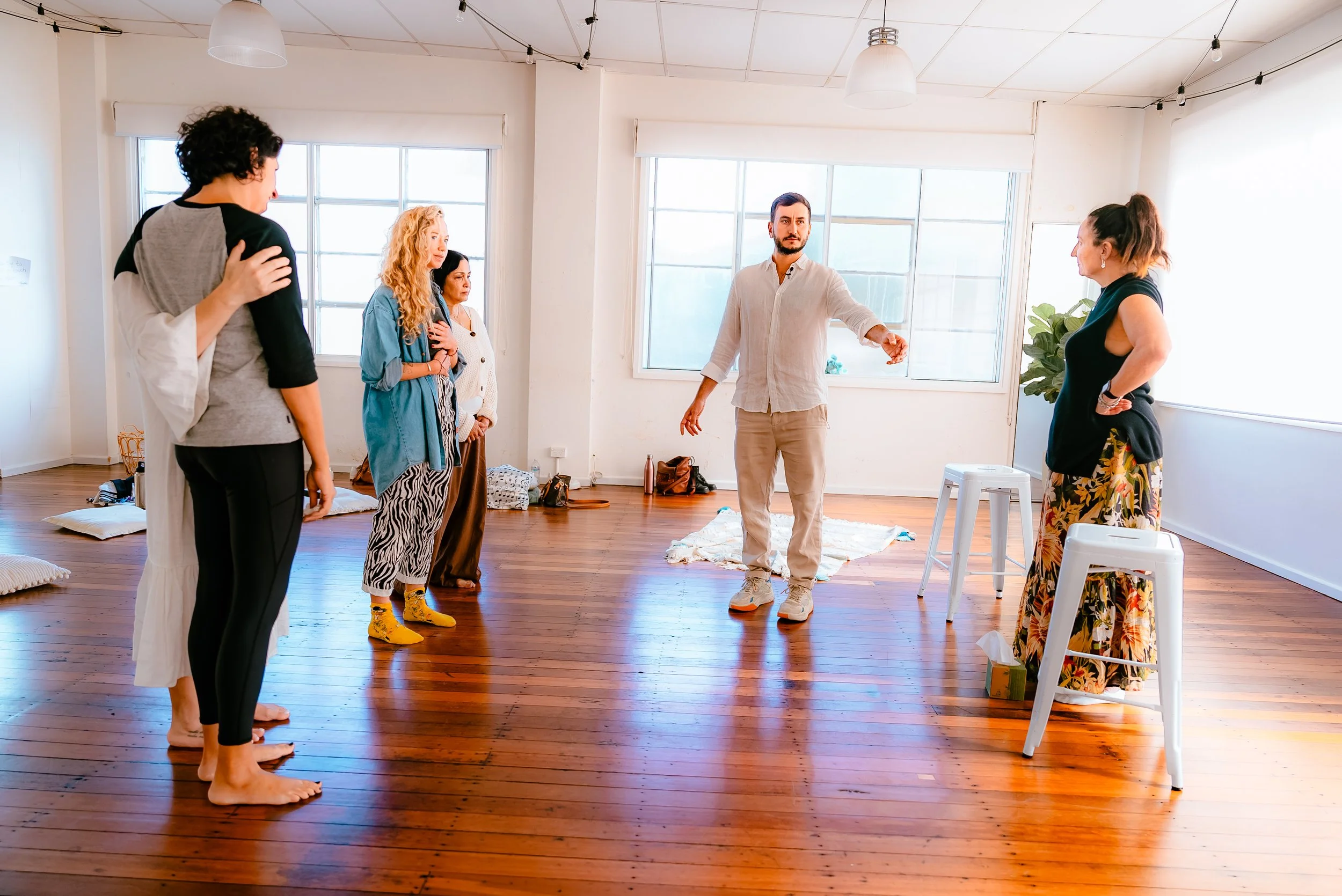 A group of six people participating in a workshop or class in a bright room with wooden floors and large windows, with some sitting on the floor and others standing.