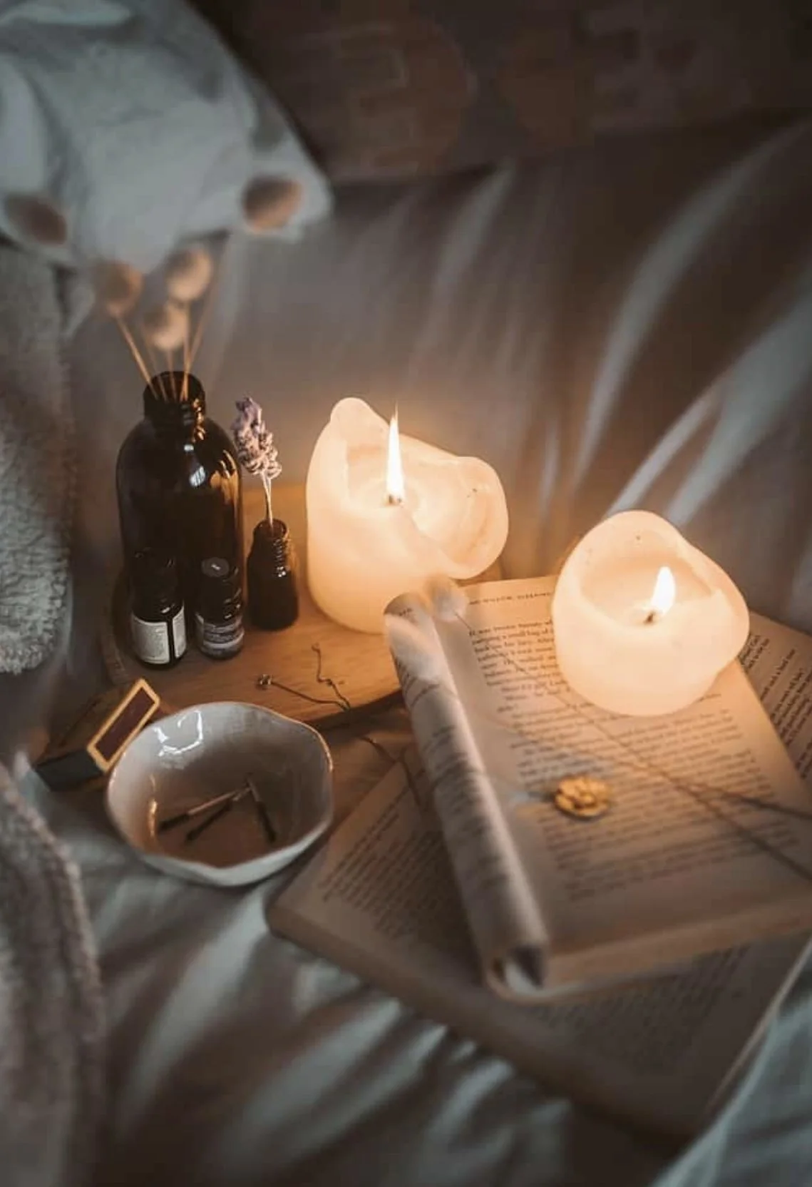 A cozy scene with two glowing salt lamp candles, an open book, a small white bowl with matches, a few essential oil bottles, dried lavender, and incense sticks on a wooden surface.