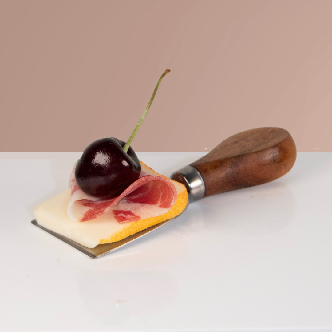 A cheese slicer with a piece of cheese, a slice of prosciutto, a cherry on top, and a cherry pitter tool on a white surface with a light pink background.