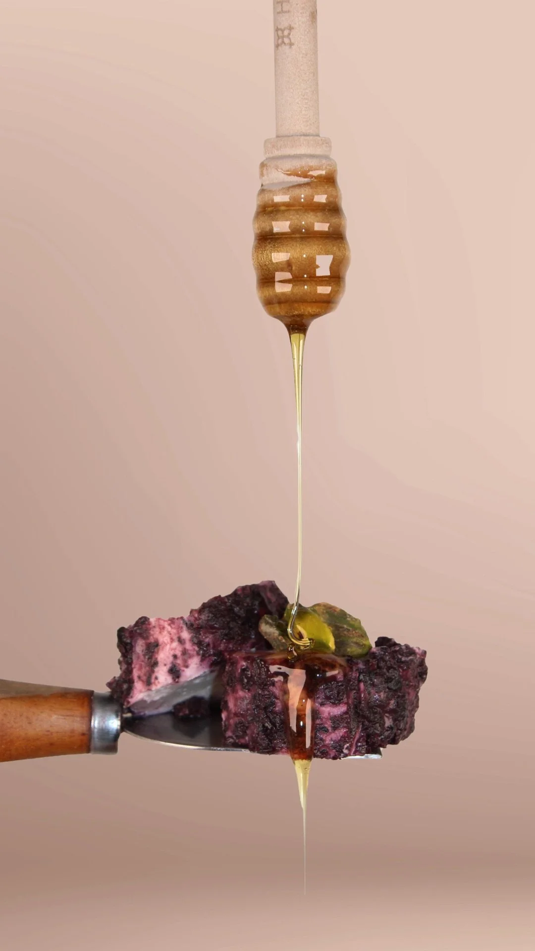 Close-up of a knife piercing a piece of blueberry cheesecake with honey dripping from a honey dipper above it.
