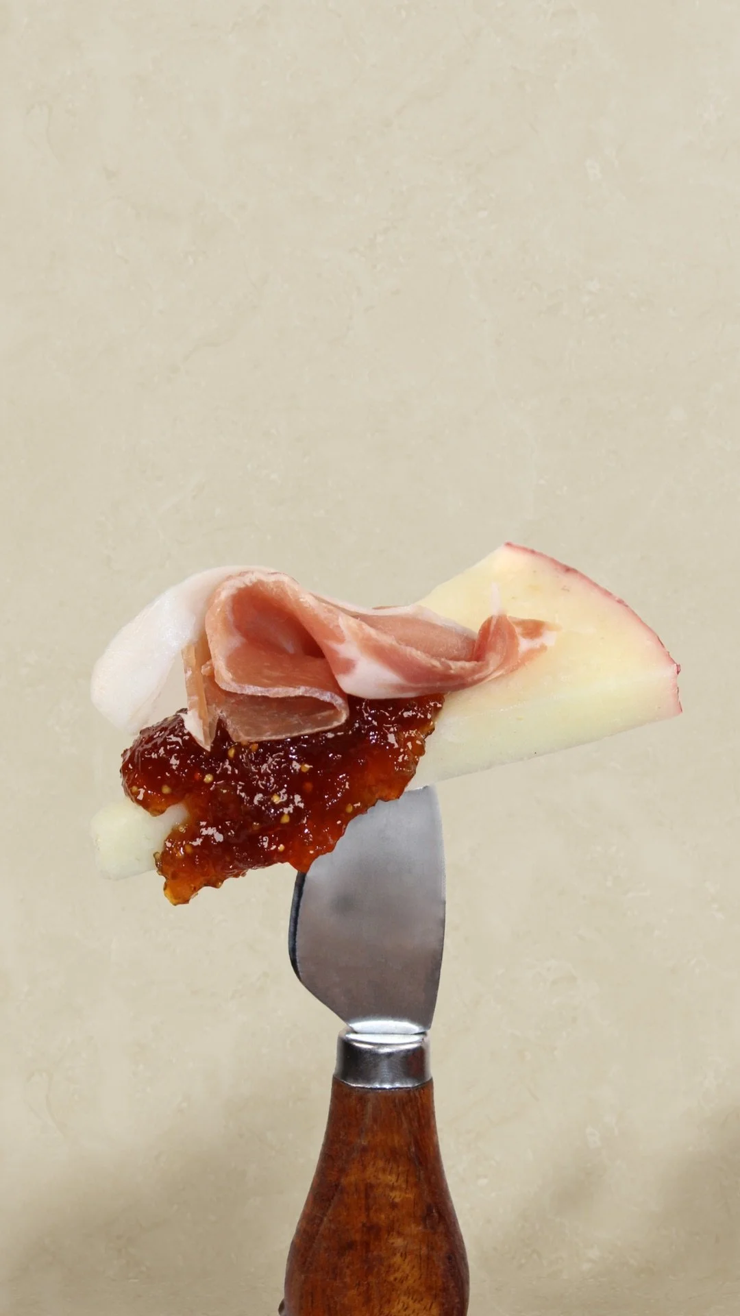 A slice of apple topped with a dollop of jam and a folded slice of prosciutto, skewered on a fork with a wooden handle against a plain background.