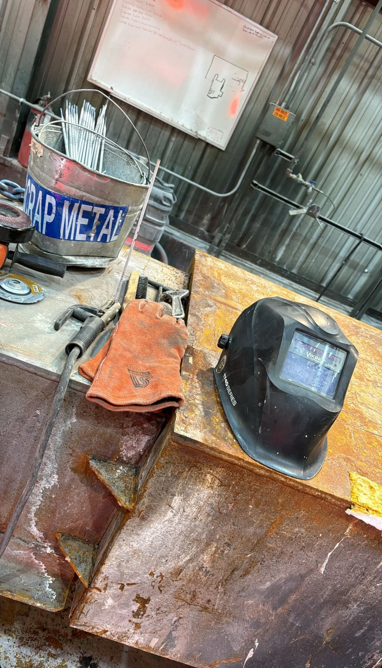 A welding workbench with a welding helmet, a worn glove, a grinder, and a bucket labeled 'Scrap Metal' filled with metal rods. In the background, a whiteboard with writing and a diagram.