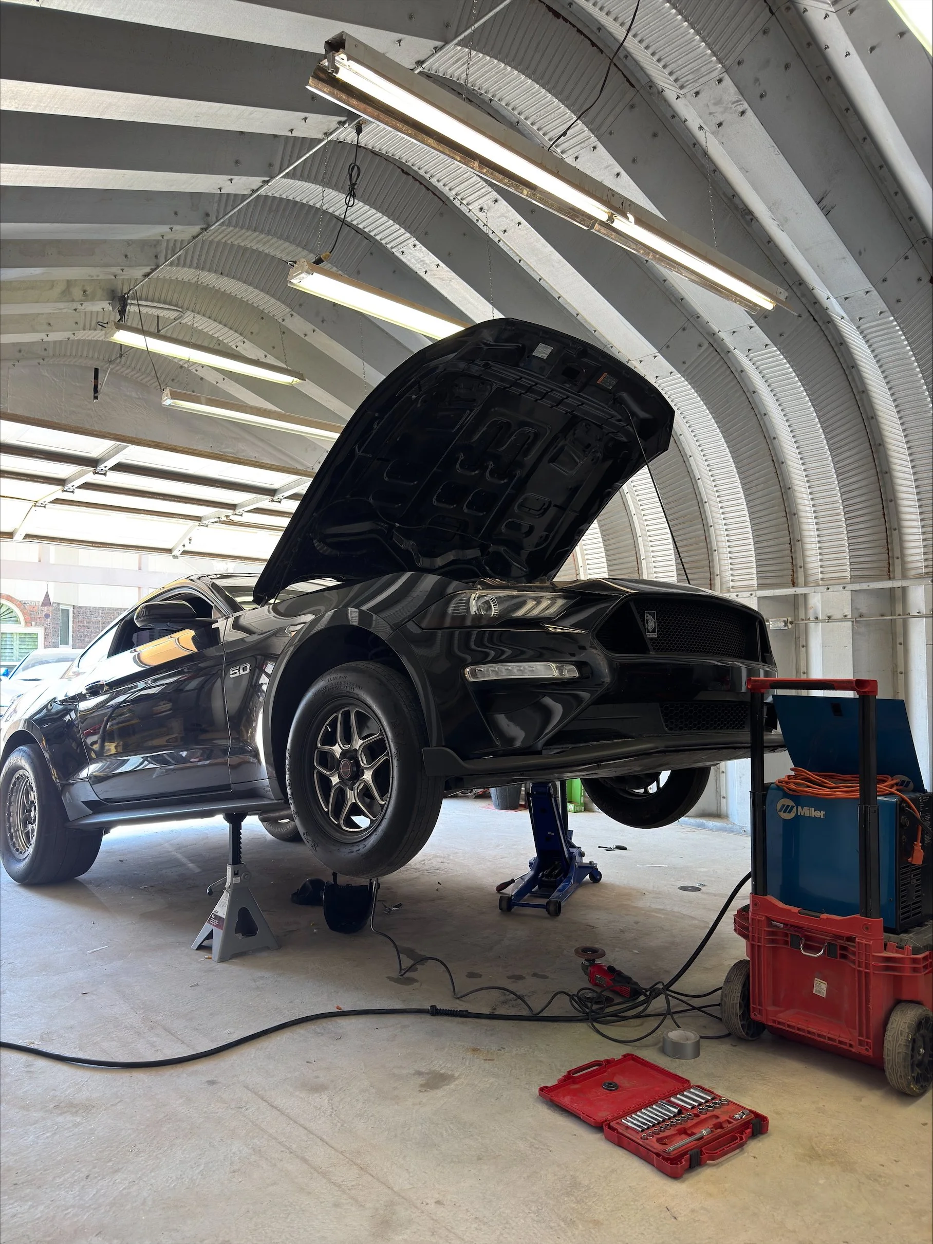 A black Ford Mustang with its hood open, raised on jack stands in a garage or workshop, with tools and equipment nearby.
