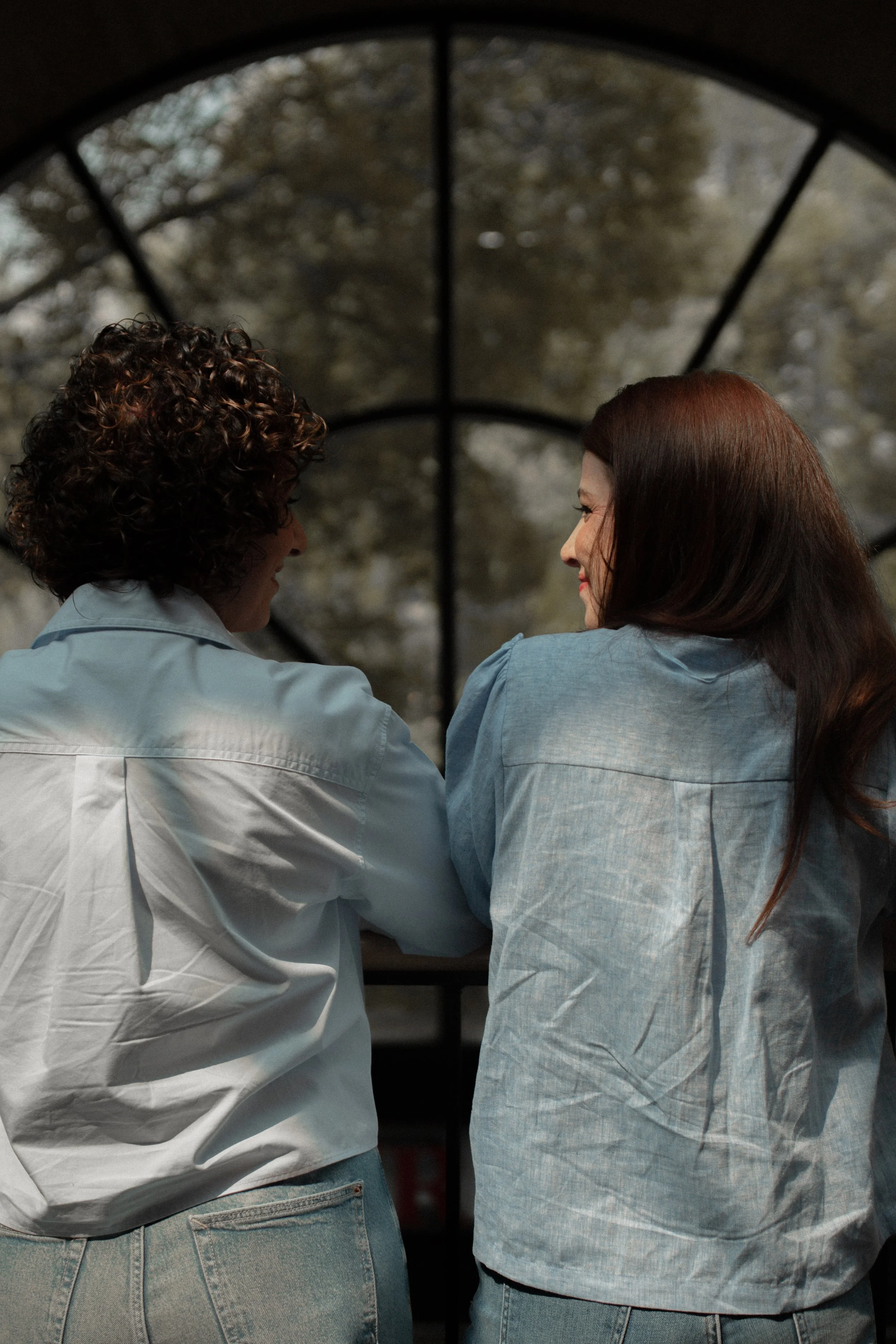 Katie and Cindy sitting side by side, smiling and talking, with large arched window behind them showing trees outside.