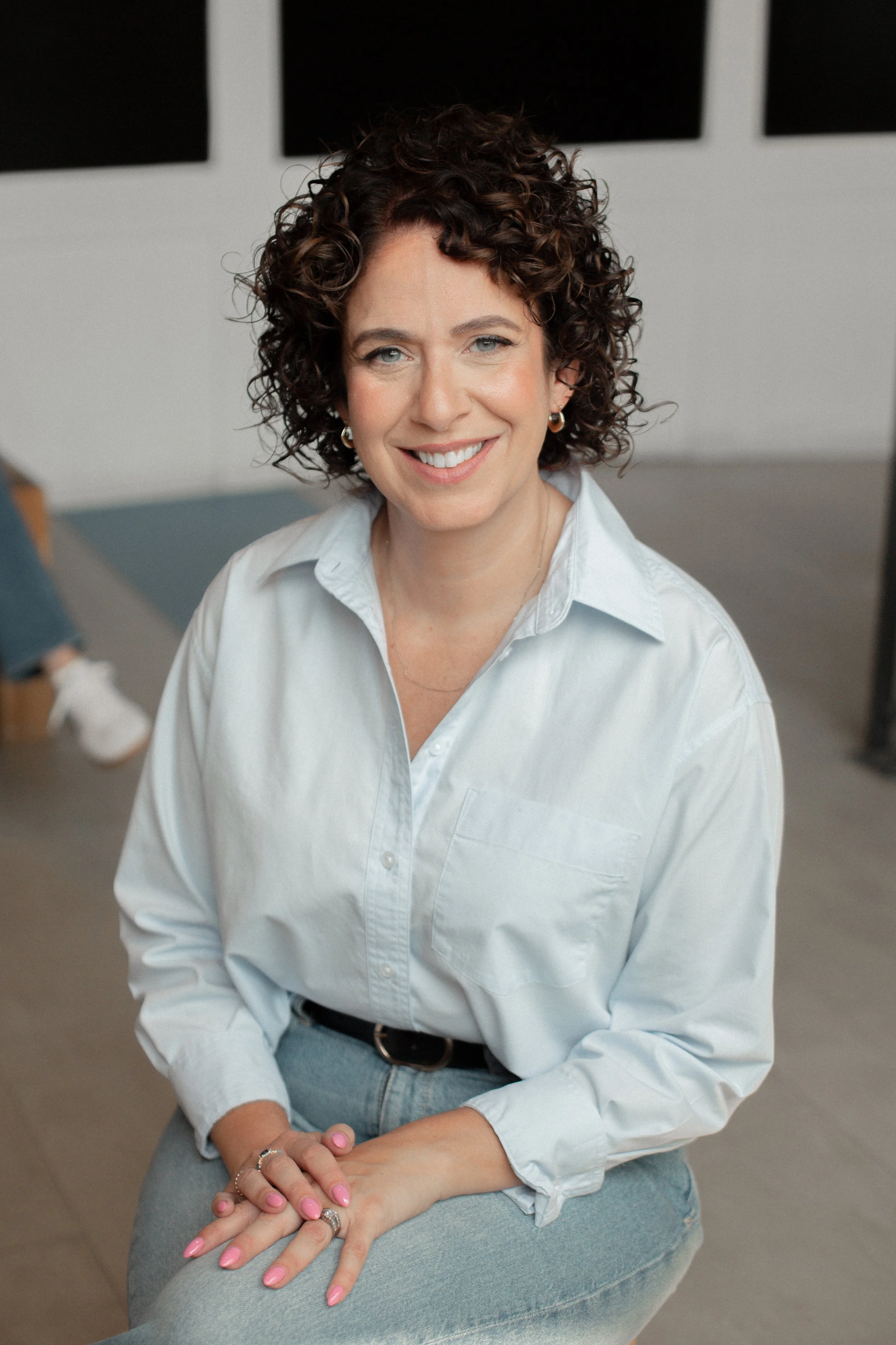 Cindy with curly brown hair, wearing a white shirt and light blue jeans, sitting indoors and smiling at the camera.