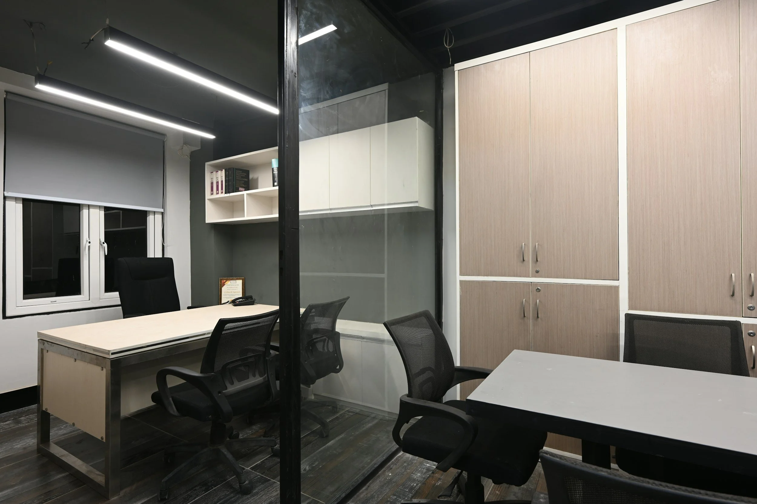 Empty office room with two desks, black office chairs, a window with a roller shade, white wall-mounted shelves, and beige cabinets.