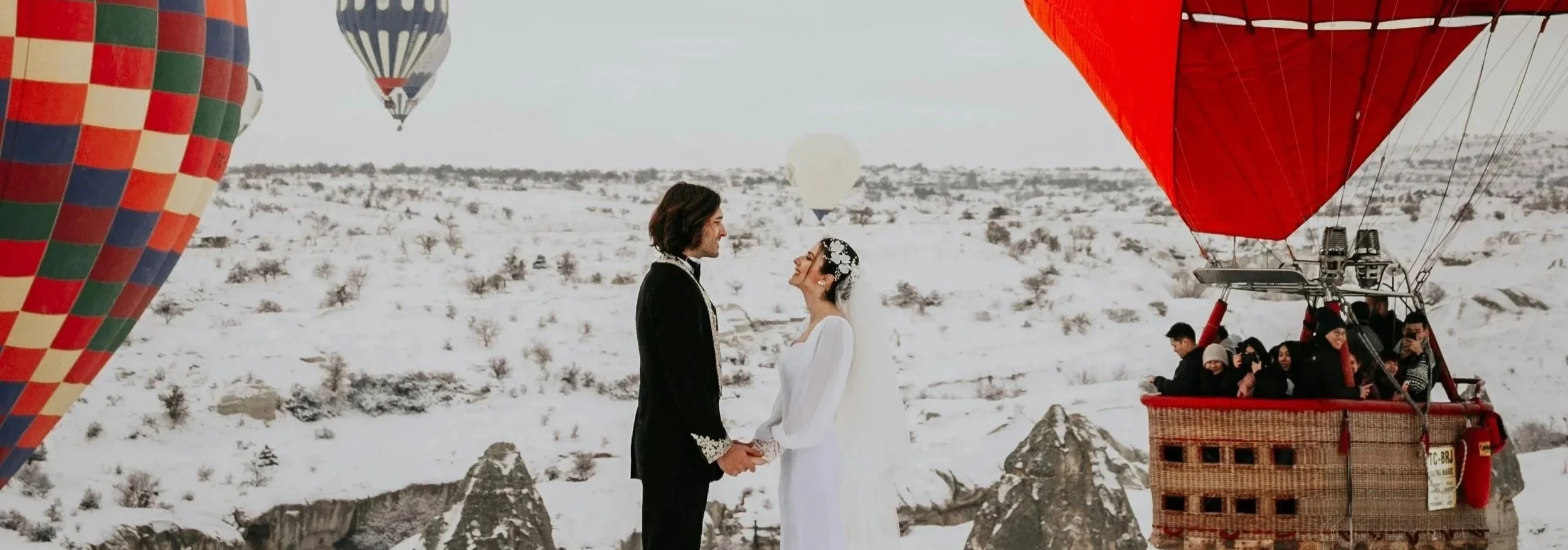 A couple in wedding attire holding hands and gazing at each other, surrounded by hot air balloons in a snowy landscape.