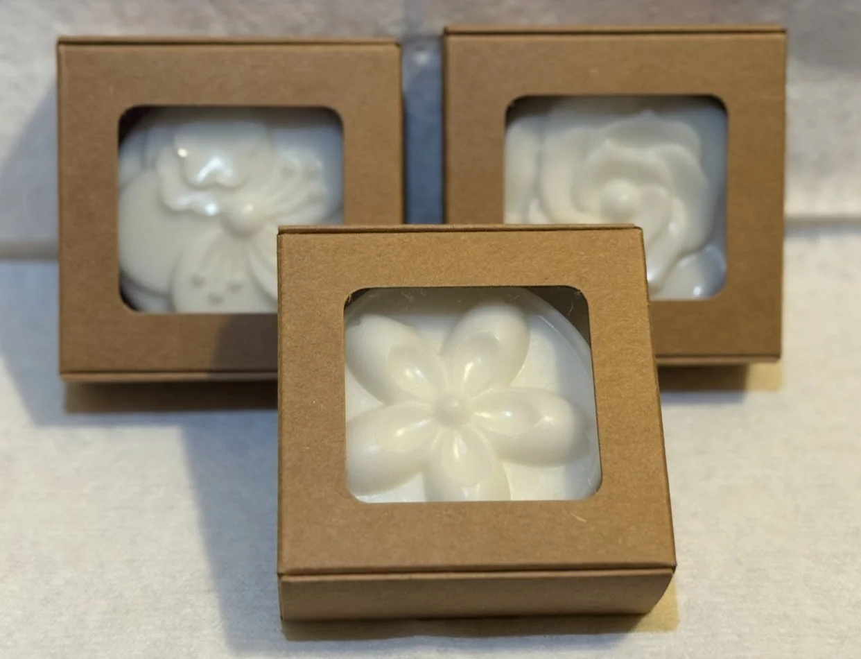 Three brown cardboard boxes with clear windows showing white soap bars with embossed flower designs.