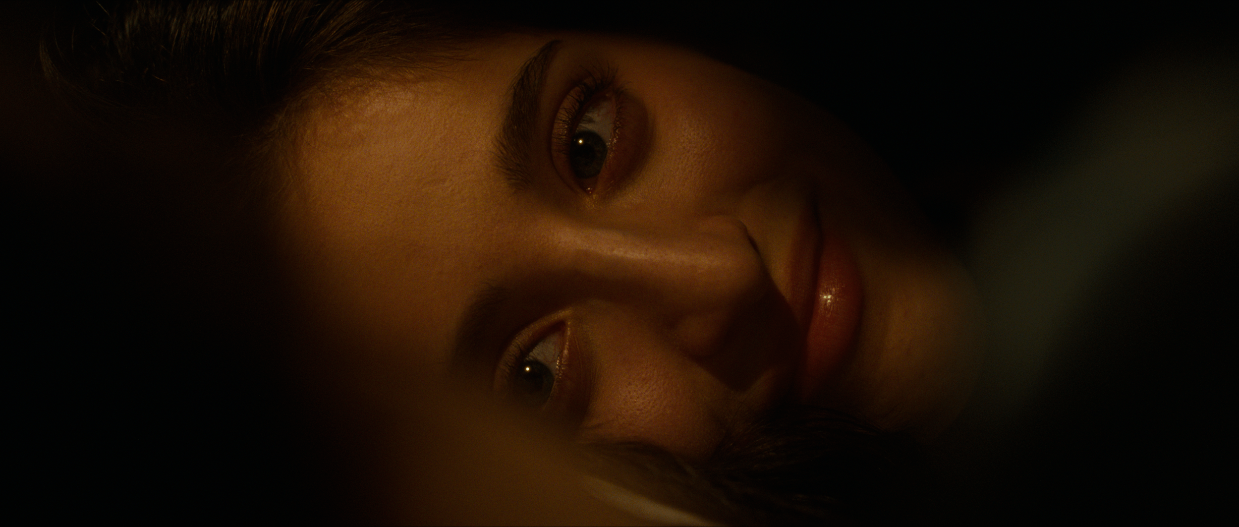Close-up of a woman's face lying on her side, with her eyes open and looking at the camera, in a dark setting.