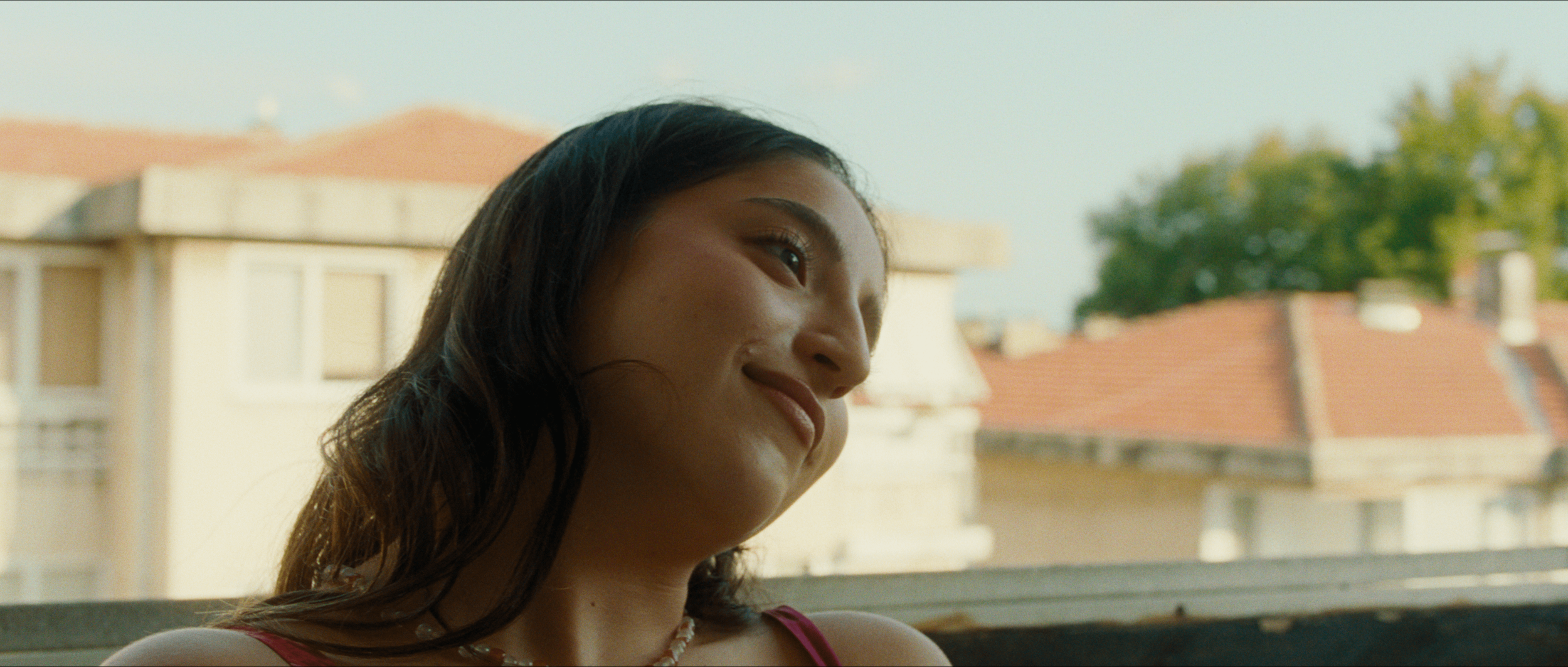 Young woman with dark hair smiling and looking to the side, outdoors with a blurred view of houses and trees in the background.