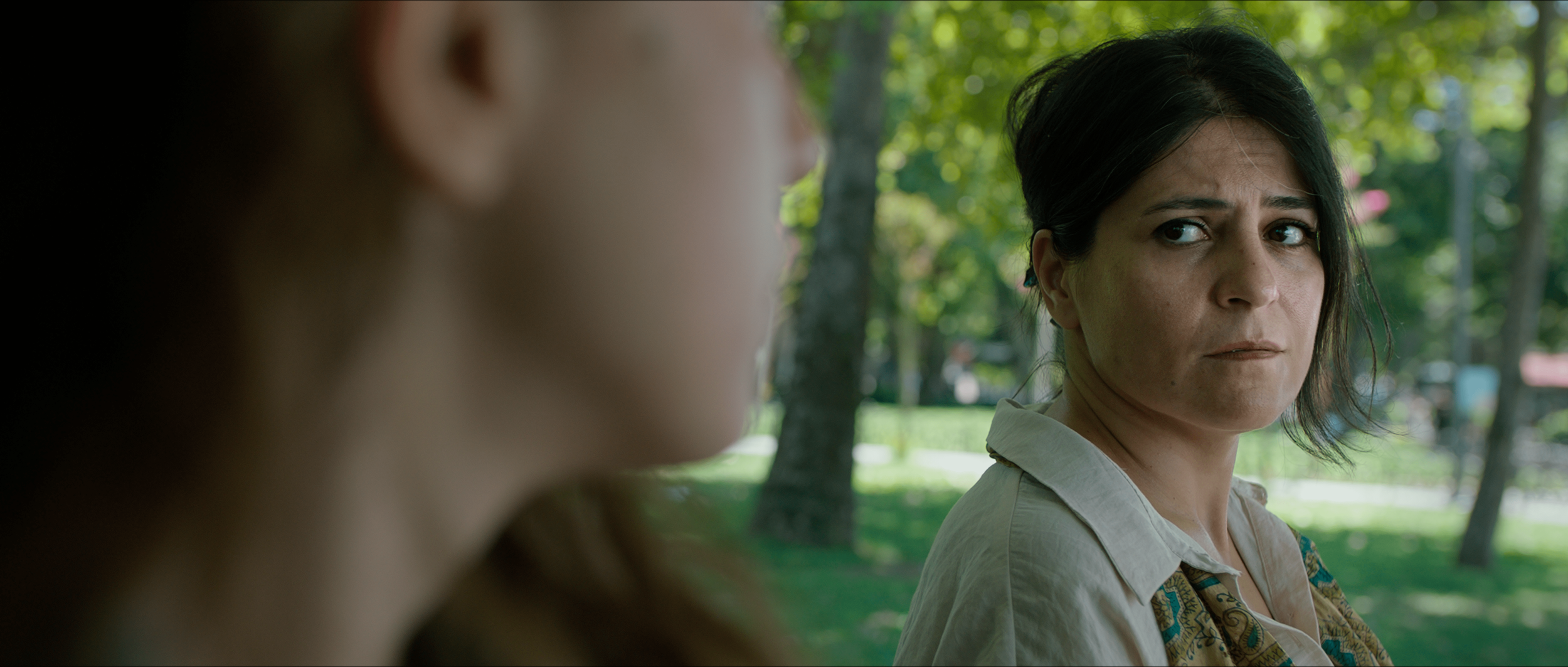 A woman with black hair and a concerned expression looking at another person who is partially visible in the foreground, in a park with green trees in the background.