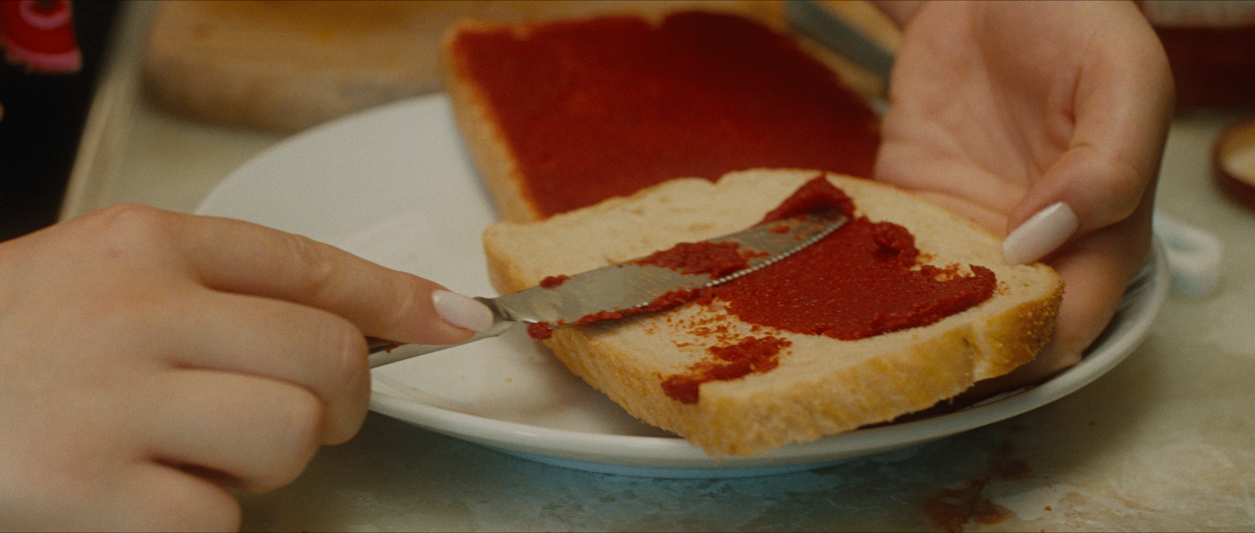 Person spreading red jam onto a slice of bread with a butter knife. Other slices of bread are on a plate in the background.