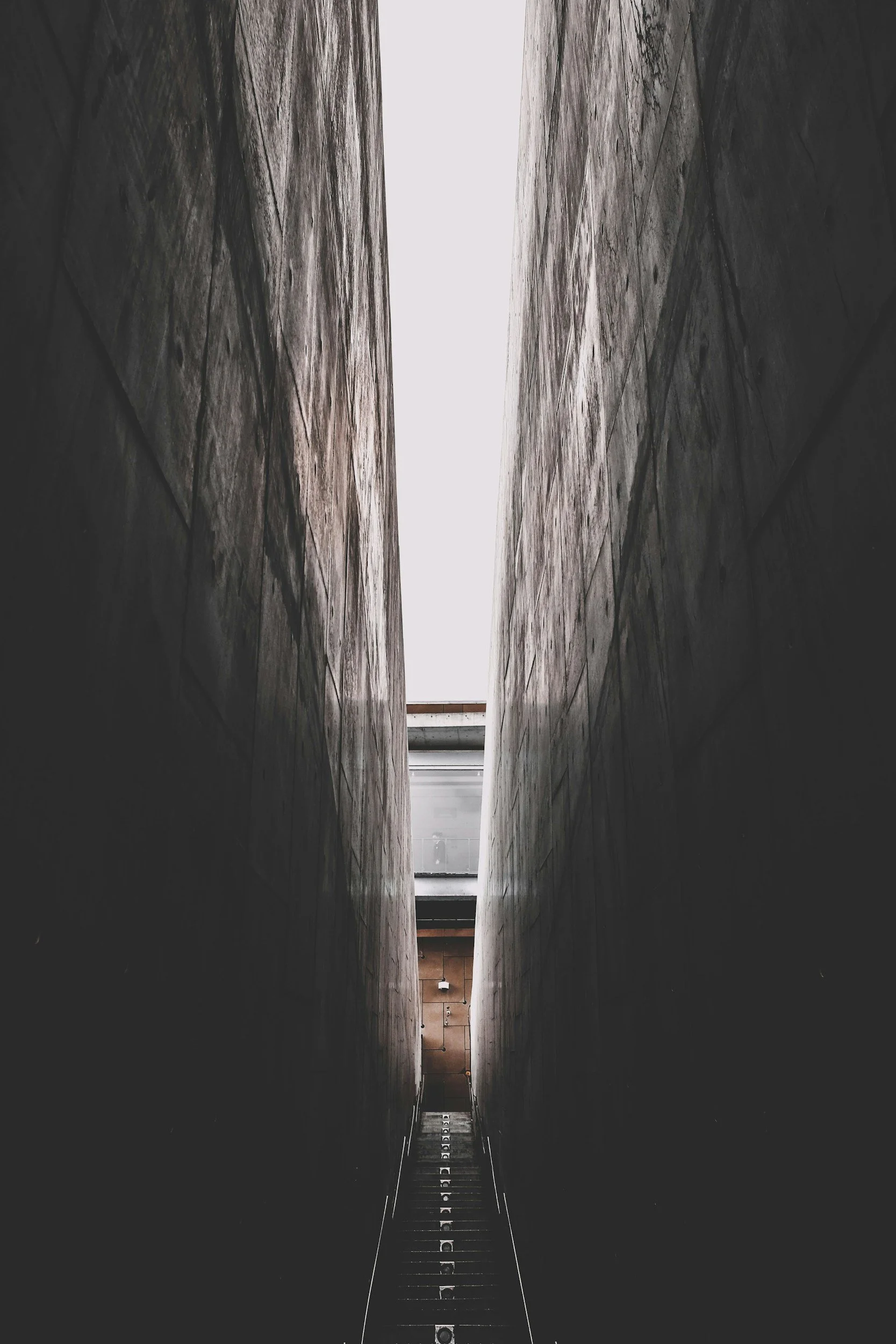 Narrow alleyway between two tall, dark stone walls, with a ladder at the bottom leading upward. Bright light at the top contrasts with shadowed walls.