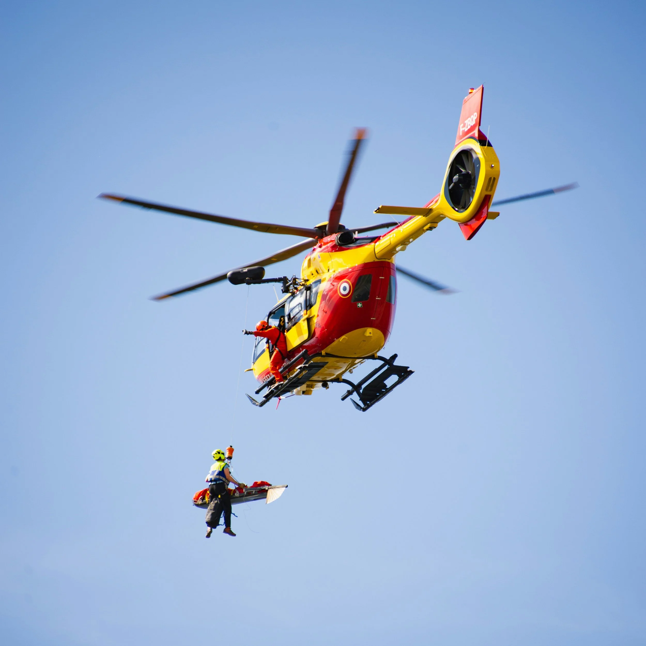 A rescue helicopter in mid-air with a crew member hanging from a rope, preparing to assist someone below against a clear blue sky.